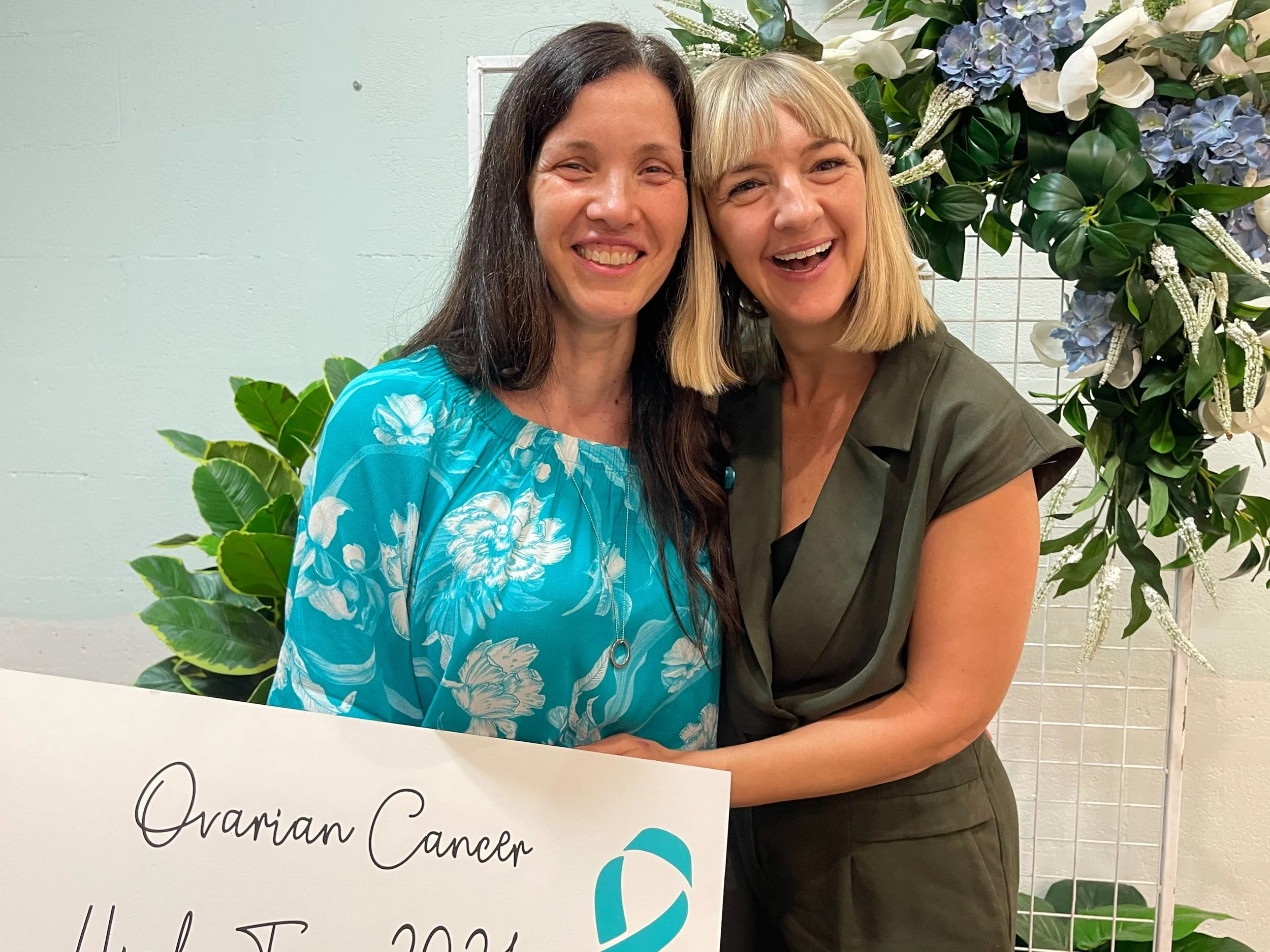 Two women smiling and hugging each other at ovarian cancer awareness event, holding a sign with text about ovarian cancer, standing in front of floral decoration.