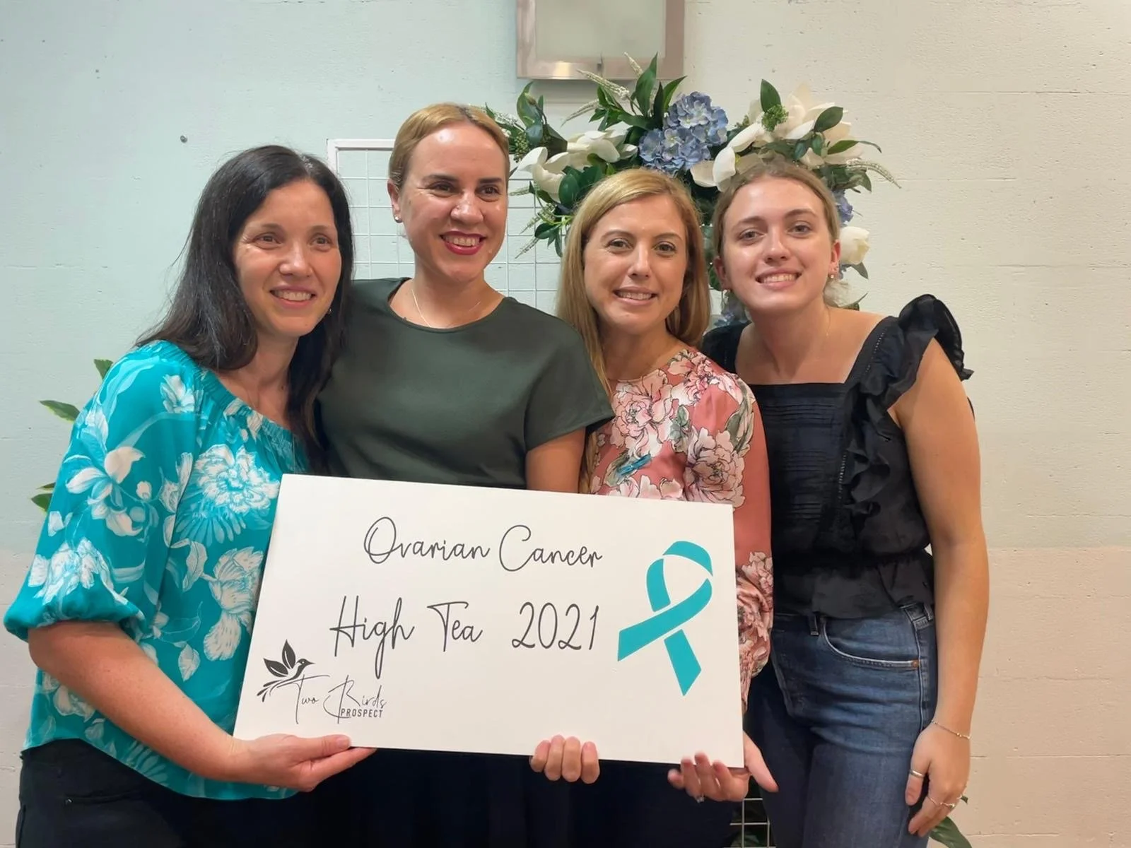 Four women smiling and standing together, holding a large white sign that reads 'Ovarian Cancer High Tea 2021' with a teal awareness ribbon and a small logo of two leaves. Behind them is a floral arrangement with white and blue flowers.