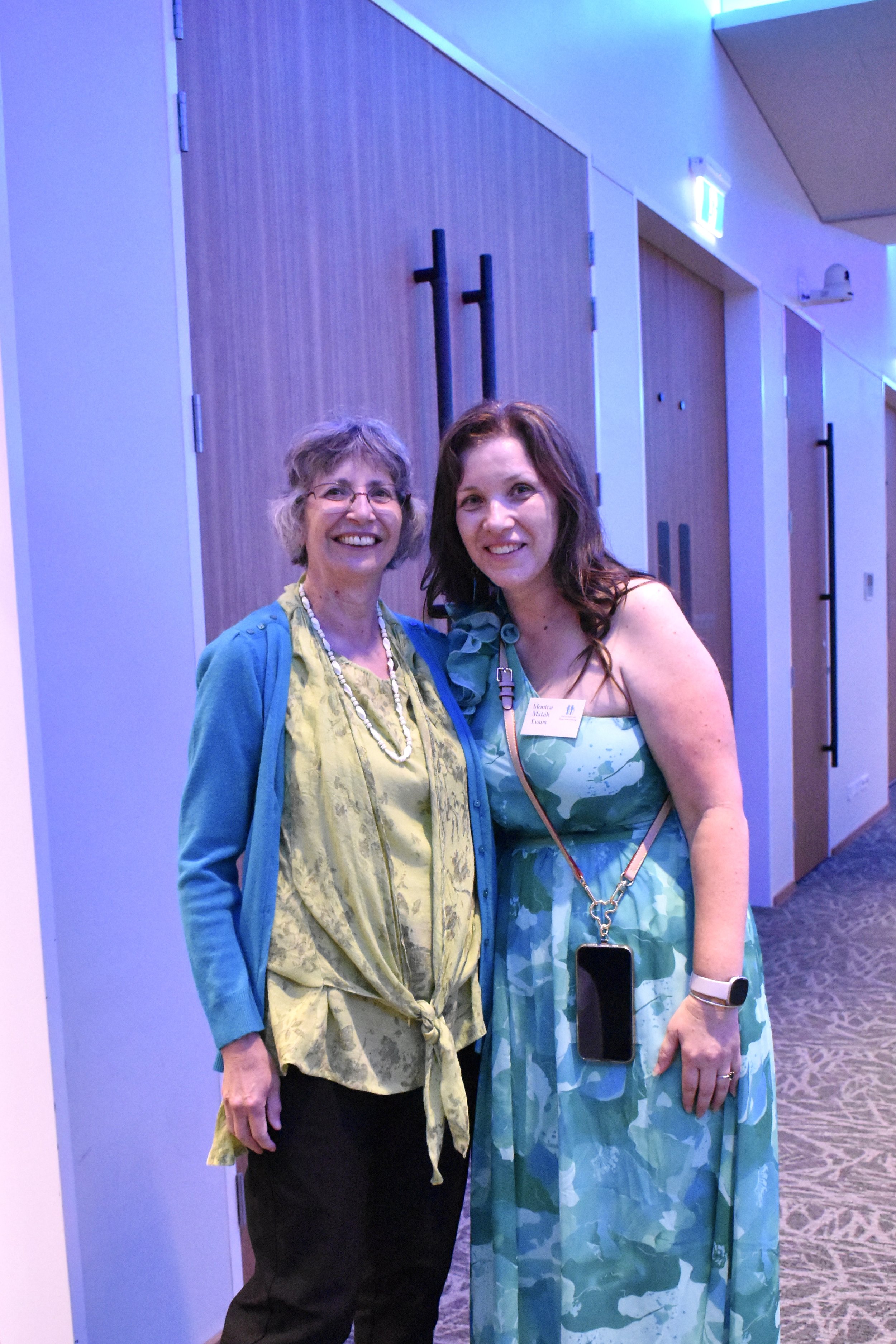 Two women standing together in a hallway smiling at the camera, at a conference or event.