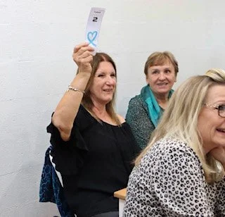 Three women sitting at a table, one woman holding up a card with a blue heart on it, in a room with white walls.