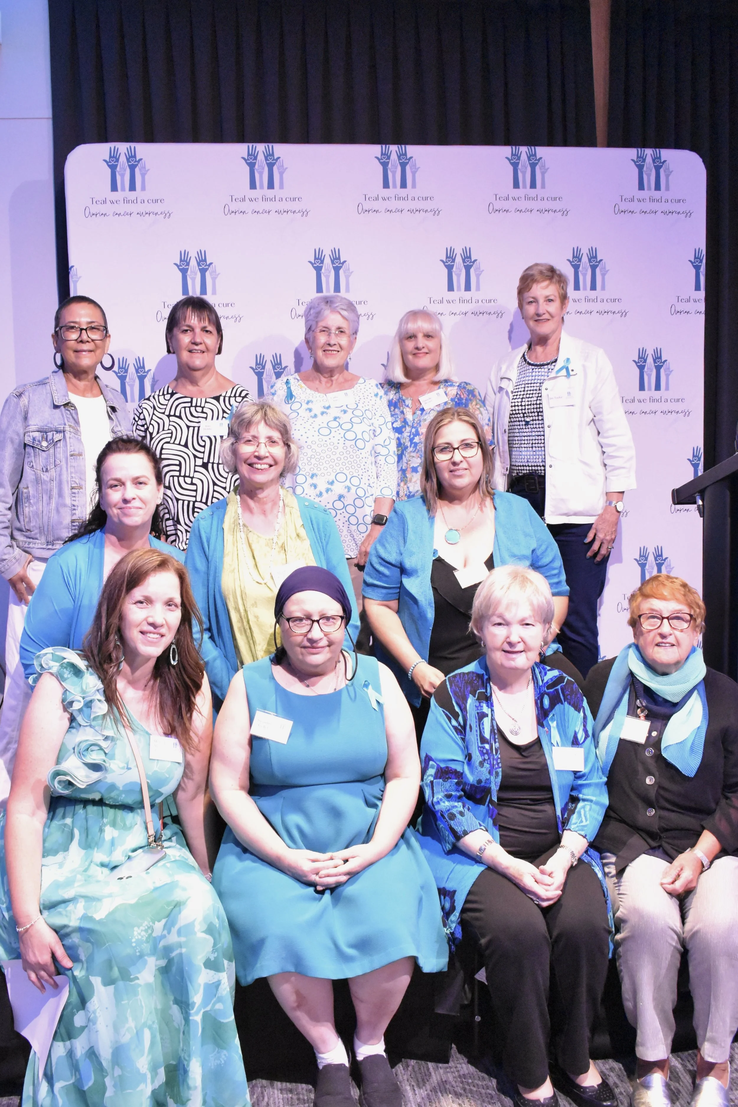 Group of women gathered at a breast cancer awareness event, with a backdrop displaying blue ribbon and hands with text reading 'Teal we find a cure, Ovarian cancer awareness.'