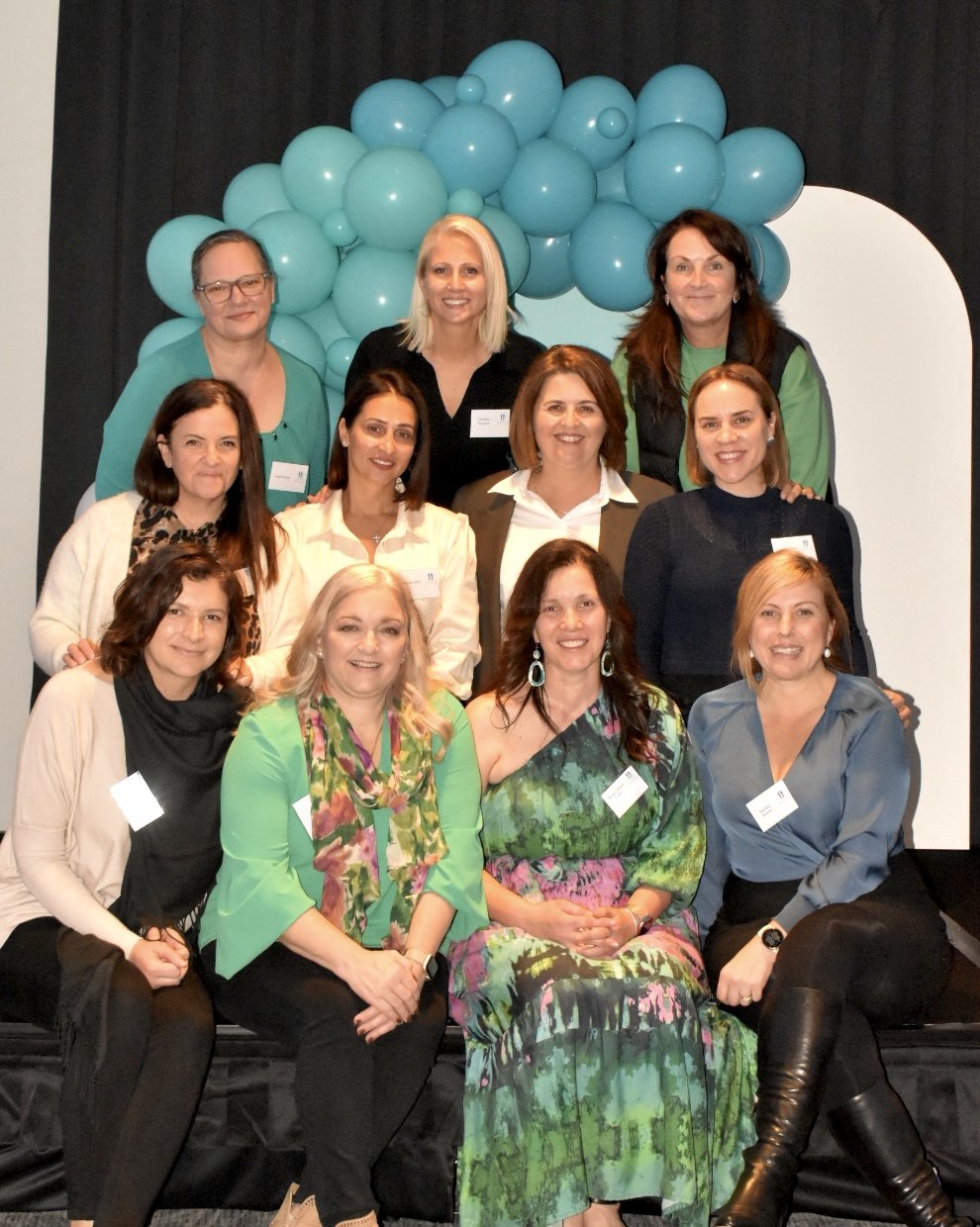 Group of women posing together on a stage with teal balloons in the background at a formal event or conference.