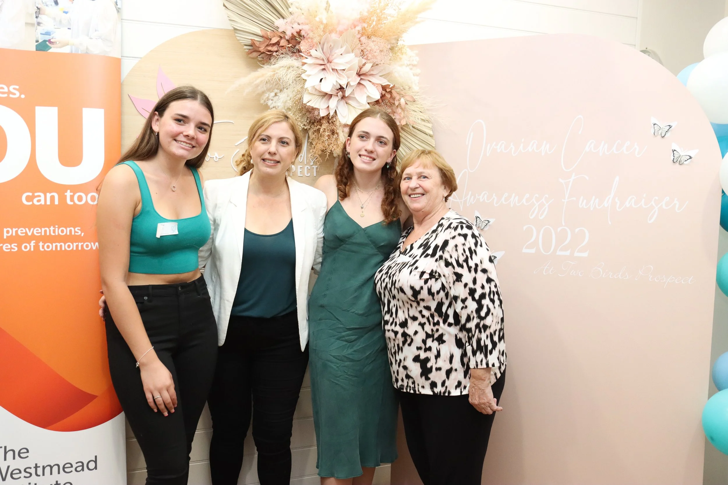 Four women standing together smiling at a cancer awareness fundraiser event, with a floral backdrop and balloons in the background.