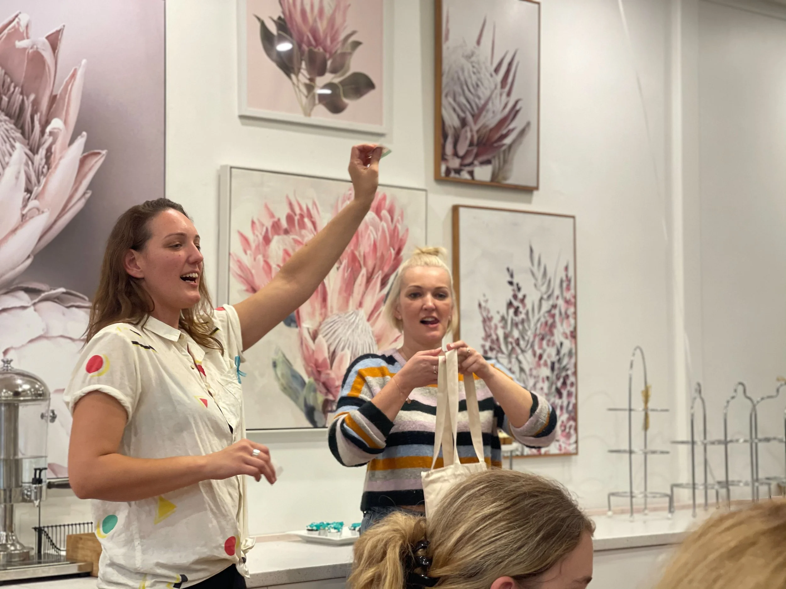 Two women standing in front of a floral art display, one is holding a gift bag and the other appears to be explaining or presenting something, with part of a person's head in the foreground.