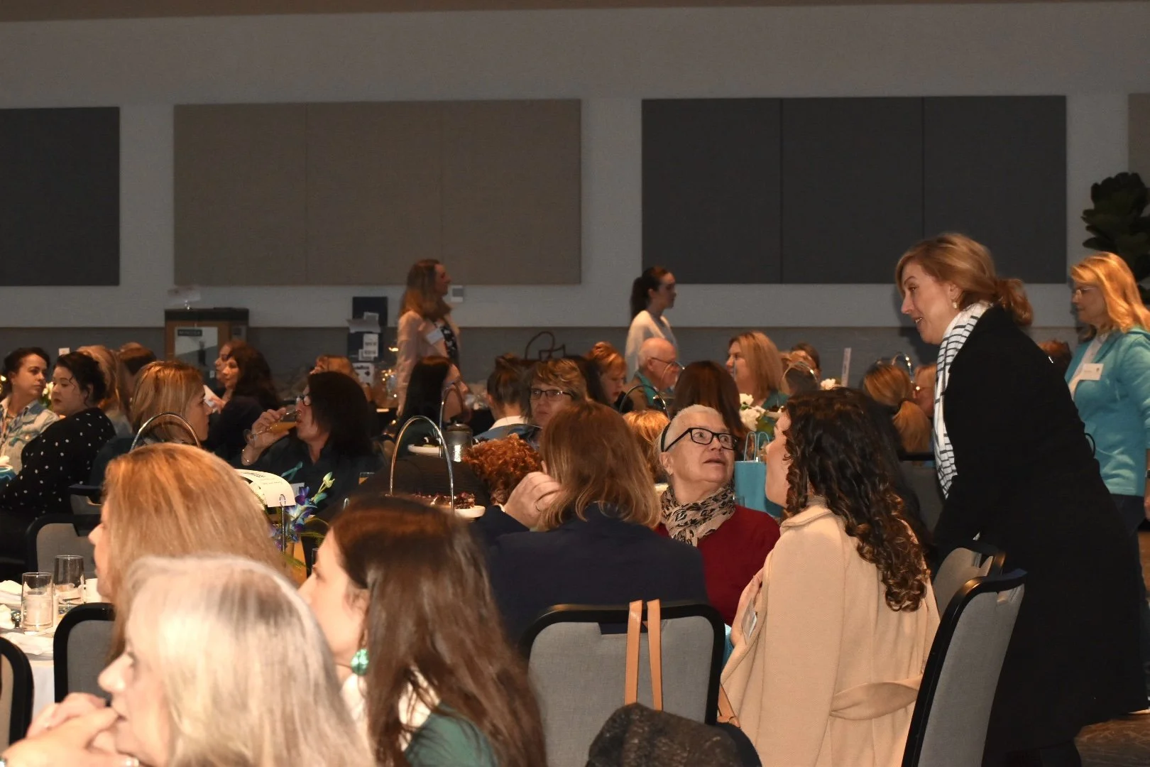 Women at a conference or banquet engaging in conversation, seated at round tables with glasses and food, with some standing and talking.
