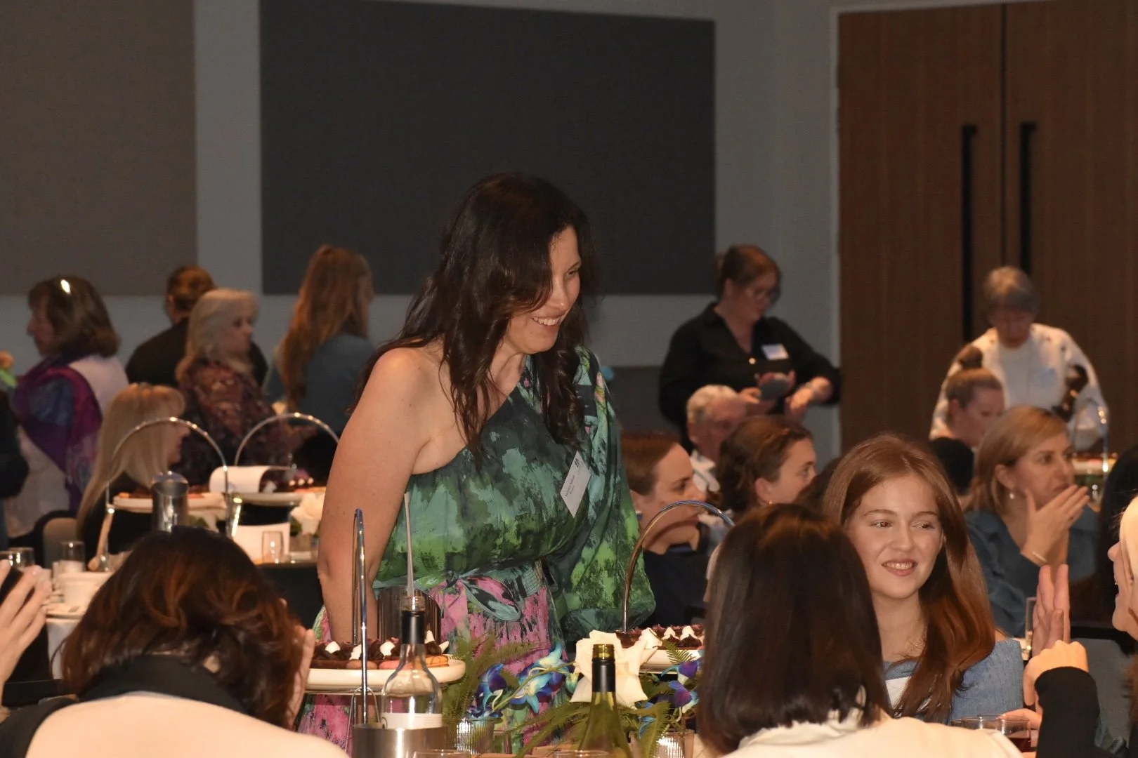 A woman with dark curly hair wearing a green and pink dress is smiling and standing at a table during a celebration or banquet. She appears to be served some food. Several women are seated around the table, some are taking photos, and others are enga
