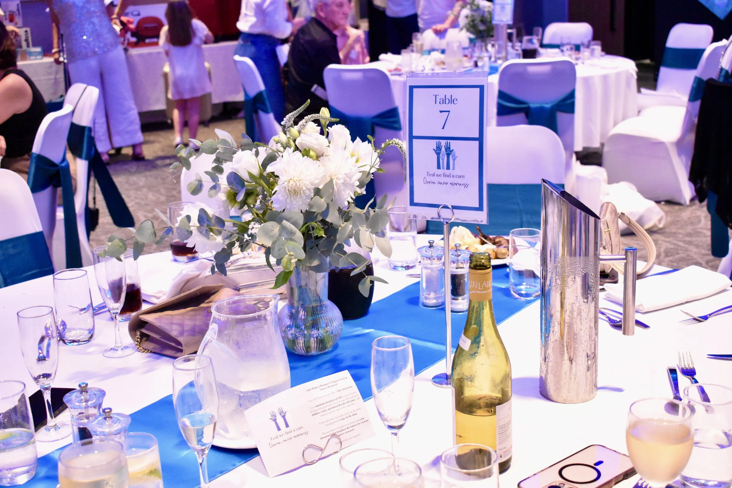 A decorated banquet table at a formal event with a floral centerpiece, glasses, a bottle of wine, and place settings, labeled as Table 7 with a sign that has a hands symbol and text about finding a cure.