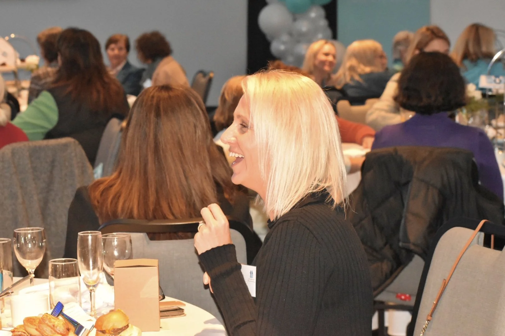 A woman with blonde hair smiling and talking at a seated gathering, with others in the background at a social event or conference.