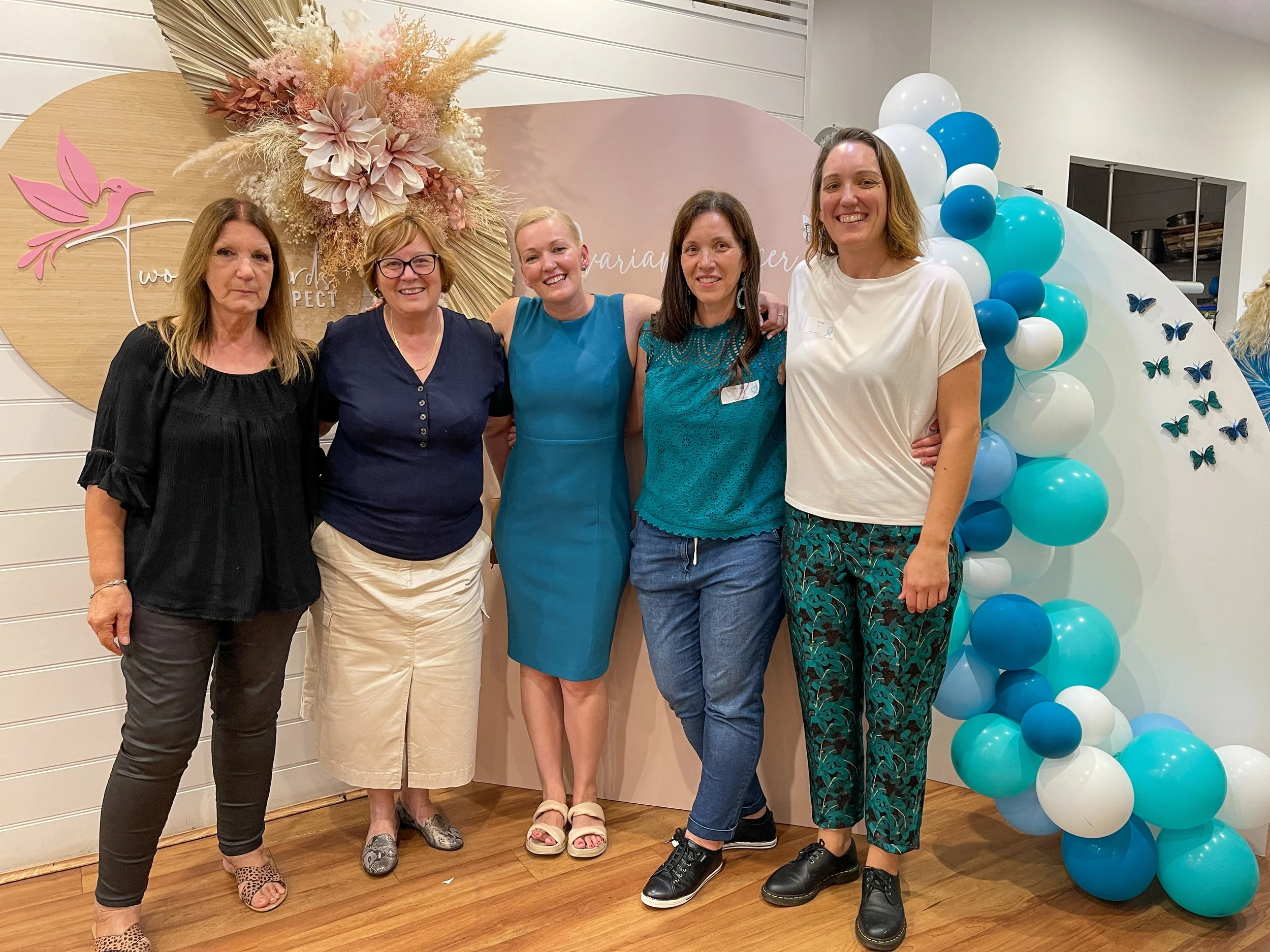 Five women standing together, smiling for a photo at a celebration or event with a decorated backdrop featuring balloons and floral arrangements.
