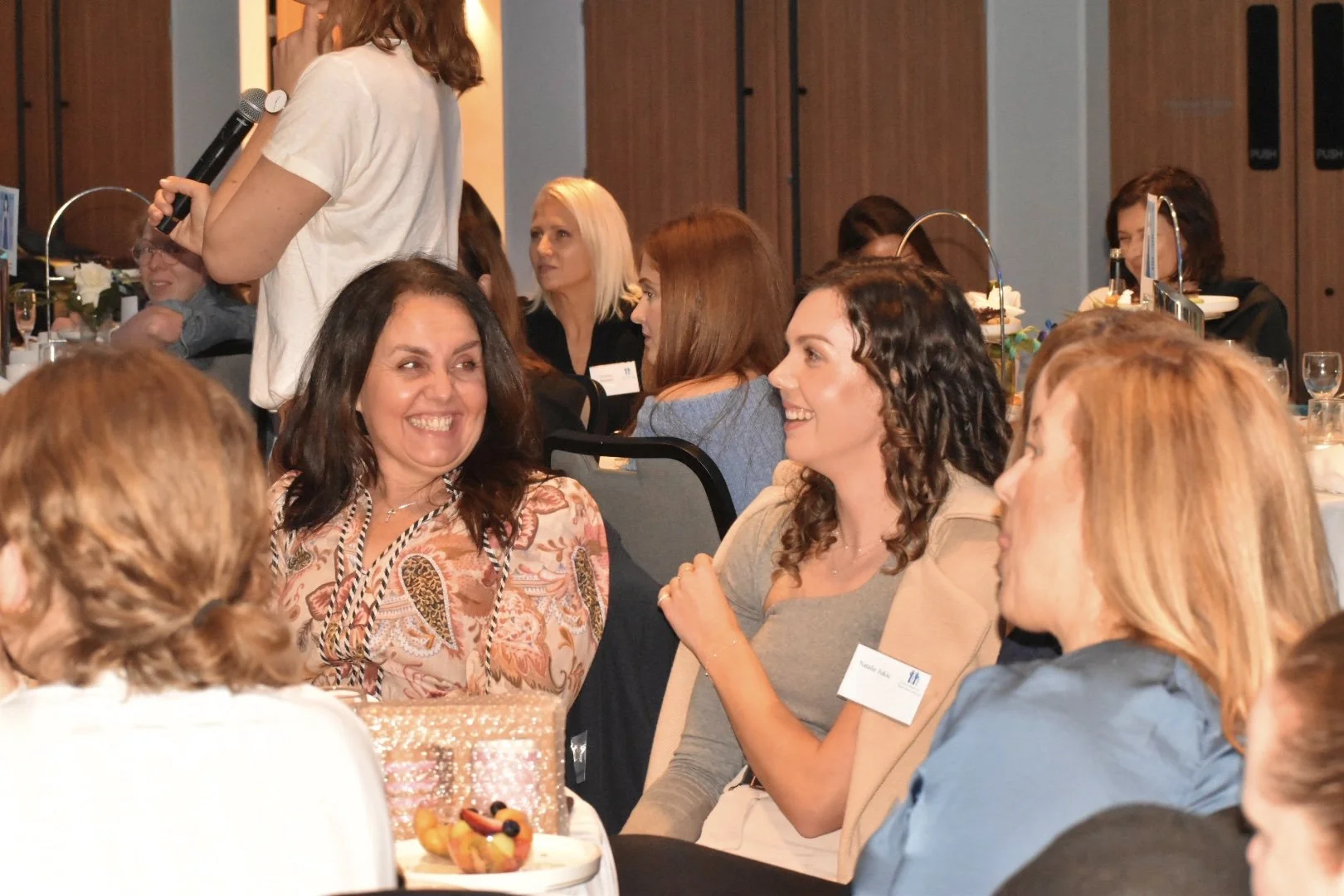 Women chatting and smiling at a conference or social event around a table with food and drinks, with one woman standing and holding a microphone.