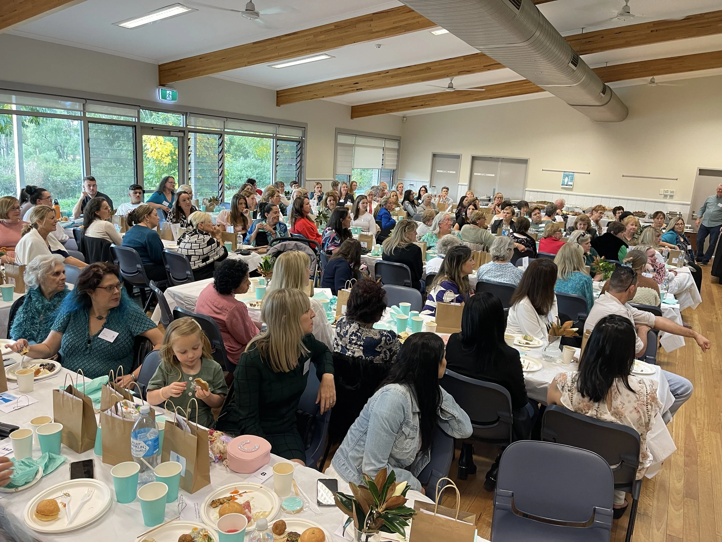 A large group of people attending a banquet or event in a spacious hall with large windows and wooden beams, seated at long tables with food, drinks, and gift bags.