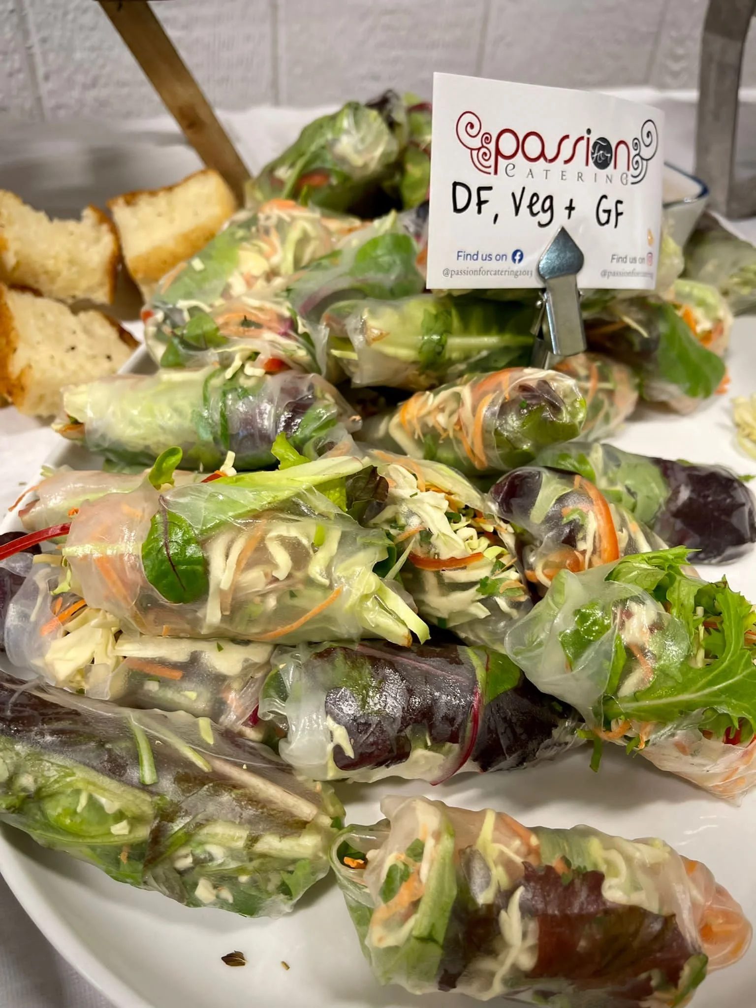 Fresh vegetable spring rolls on a white plate with a sign indicating they are vegan and gluten-free, accompanied by pieces of bread in the background.