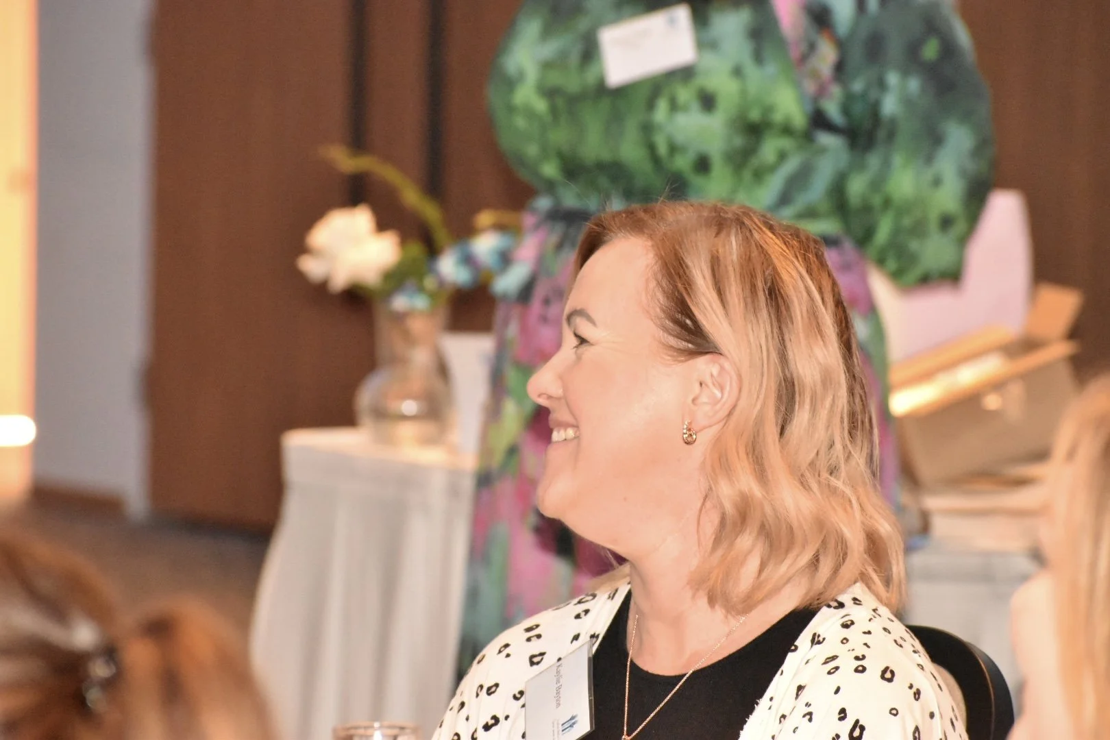 Profile of a woman with shoulder-length strawberry blonde hair smiling, wearing a white and black spotted top, small hoop earrings, and a gold necklace, sitting at a table during an indoor event.