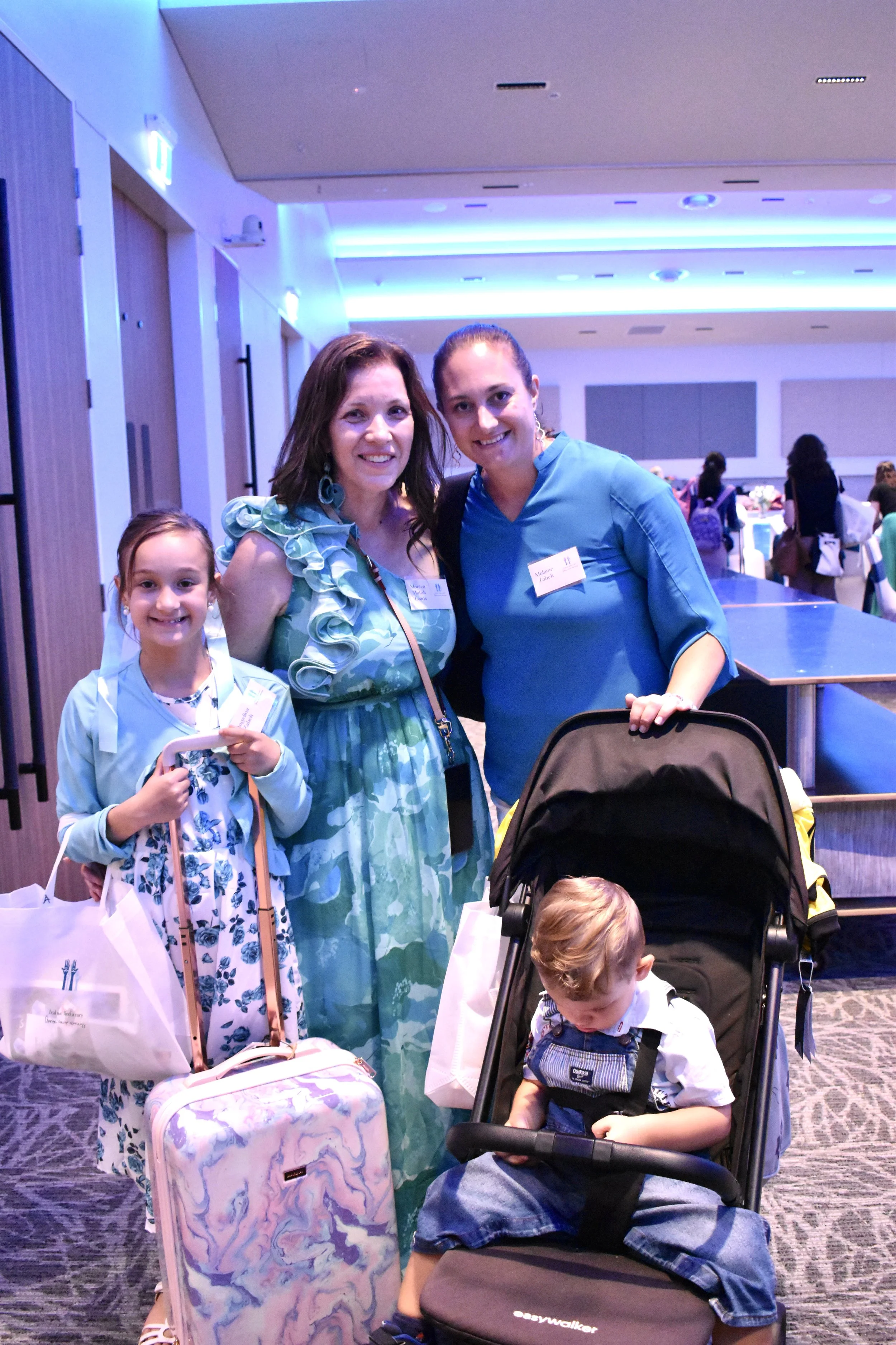 A group of four people, including two women, a young girl, and a boy in a stroller, at an indoor event or conference, with others in the background.