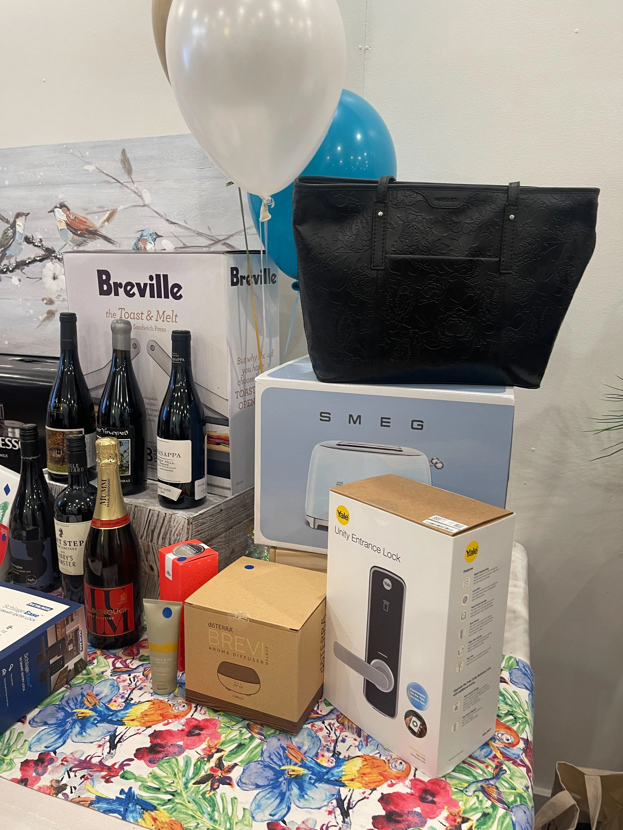 Table with various products including bottles of wine, an aroma diffuser, a smart lock, and a black tote bag. There are balloons and boxed appliances in the background.