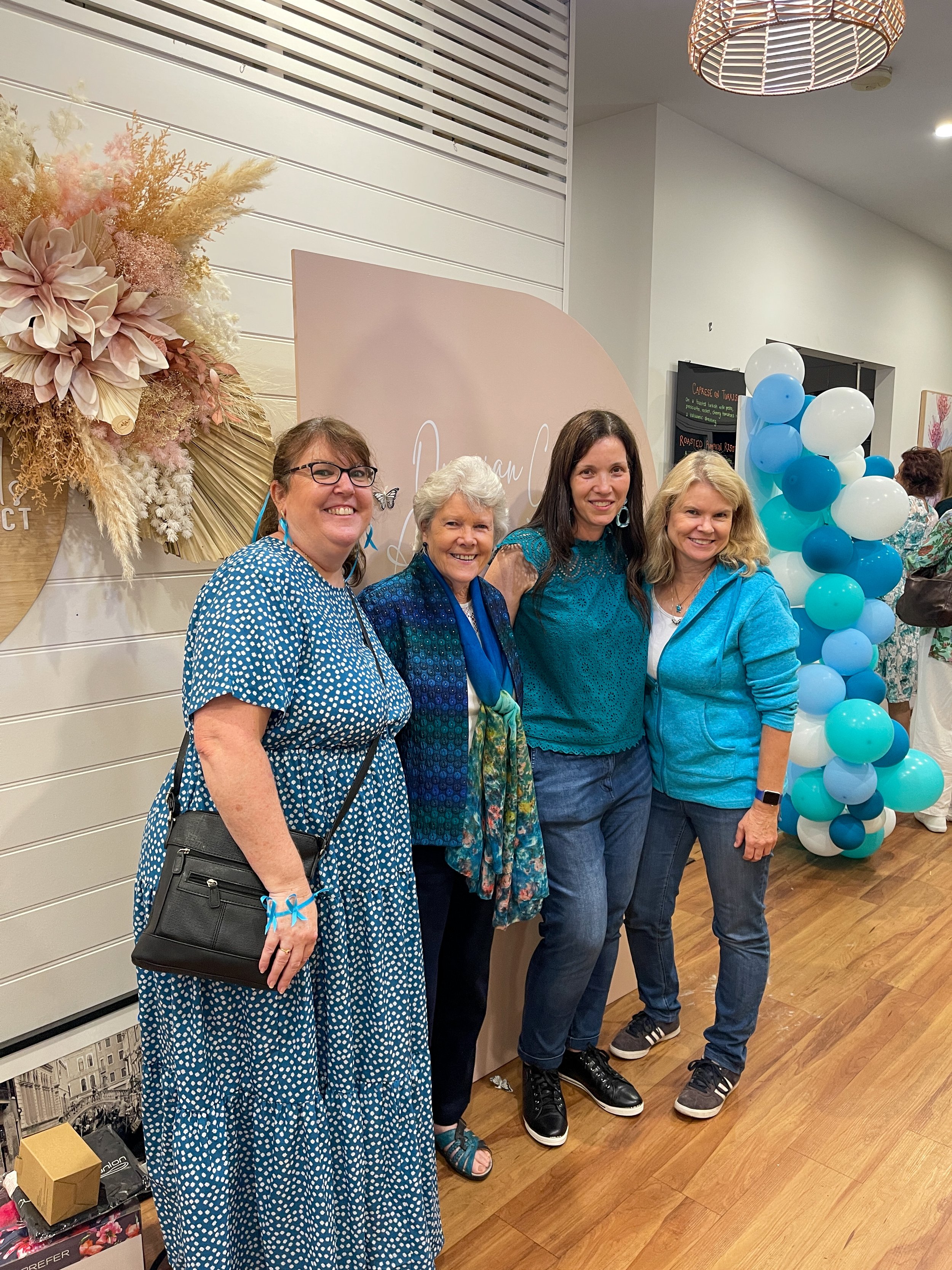 Four women standing together at an indoor event with a pink sign reading 'Dream' and pastel-colored balloon decorations in the background.