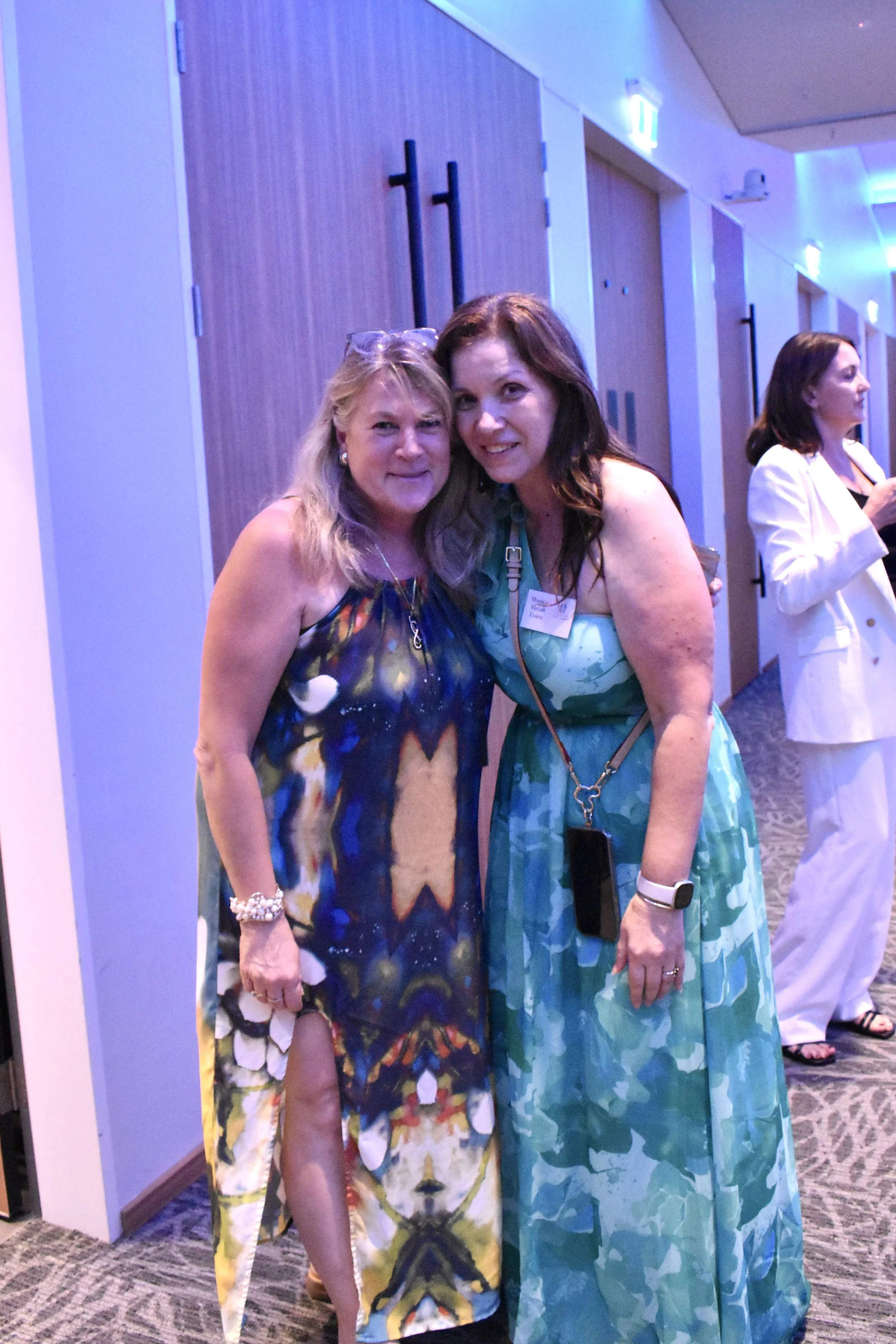 Two women standing close together at an indoor event, smiling for the camera. The woman on the left has blonde hair and is wearing a colorful, sleeveless, high-slit dress. The woman on the right has brown hair, is wearing a teal pattern maxi dress, a