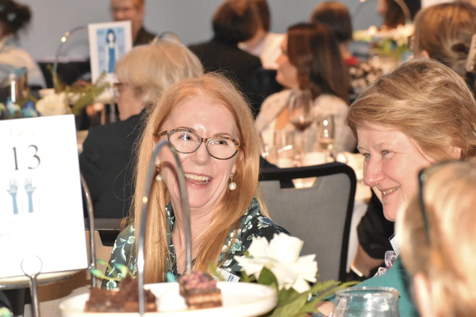 Women smiling and engaging in conversation at a formal event with a table set for a meal, flowers, and a table number card.