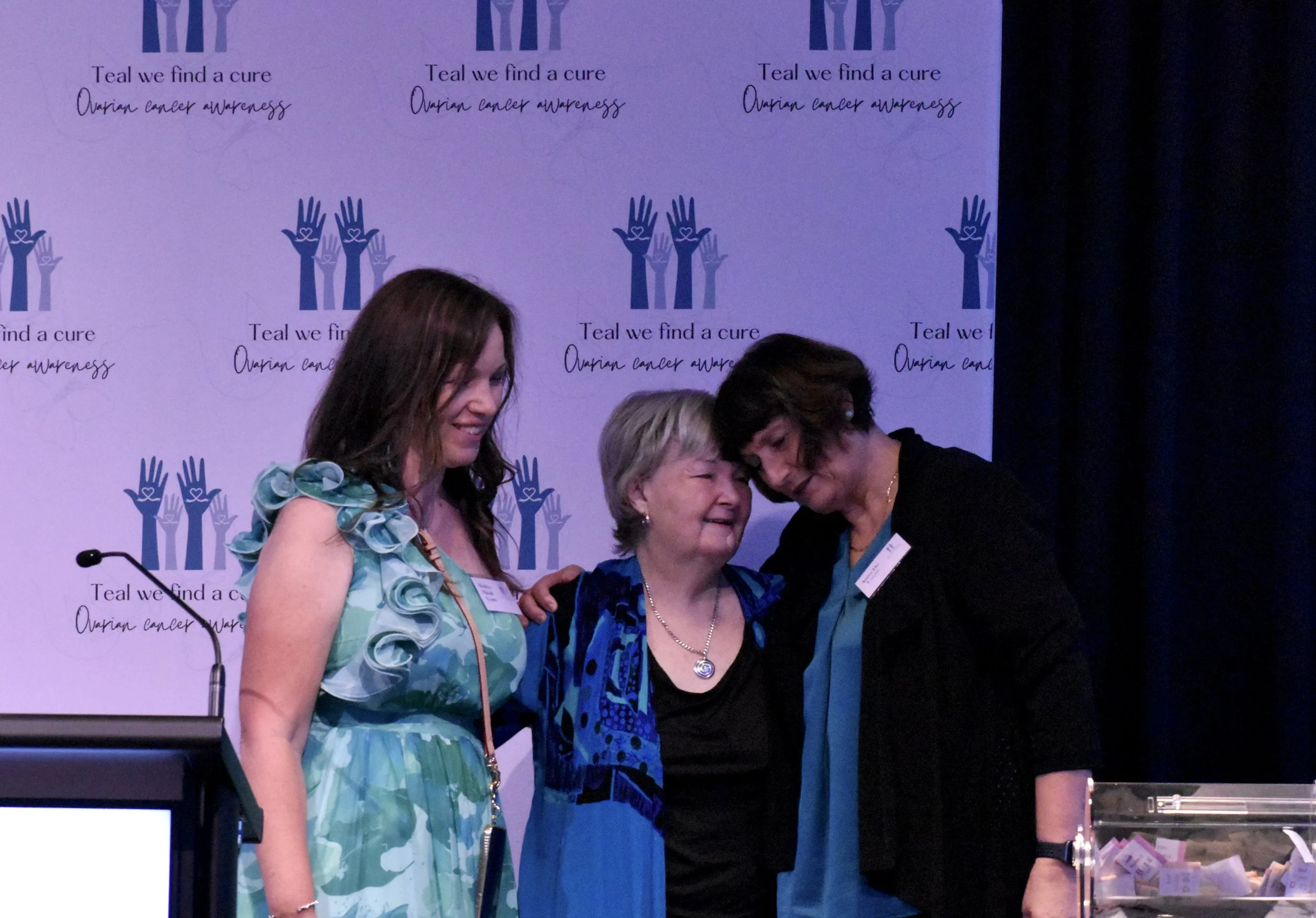 Three women at an ovarian cancer awareness event, standing in front of a backdrop with text and logo, clasping hands and smiling.