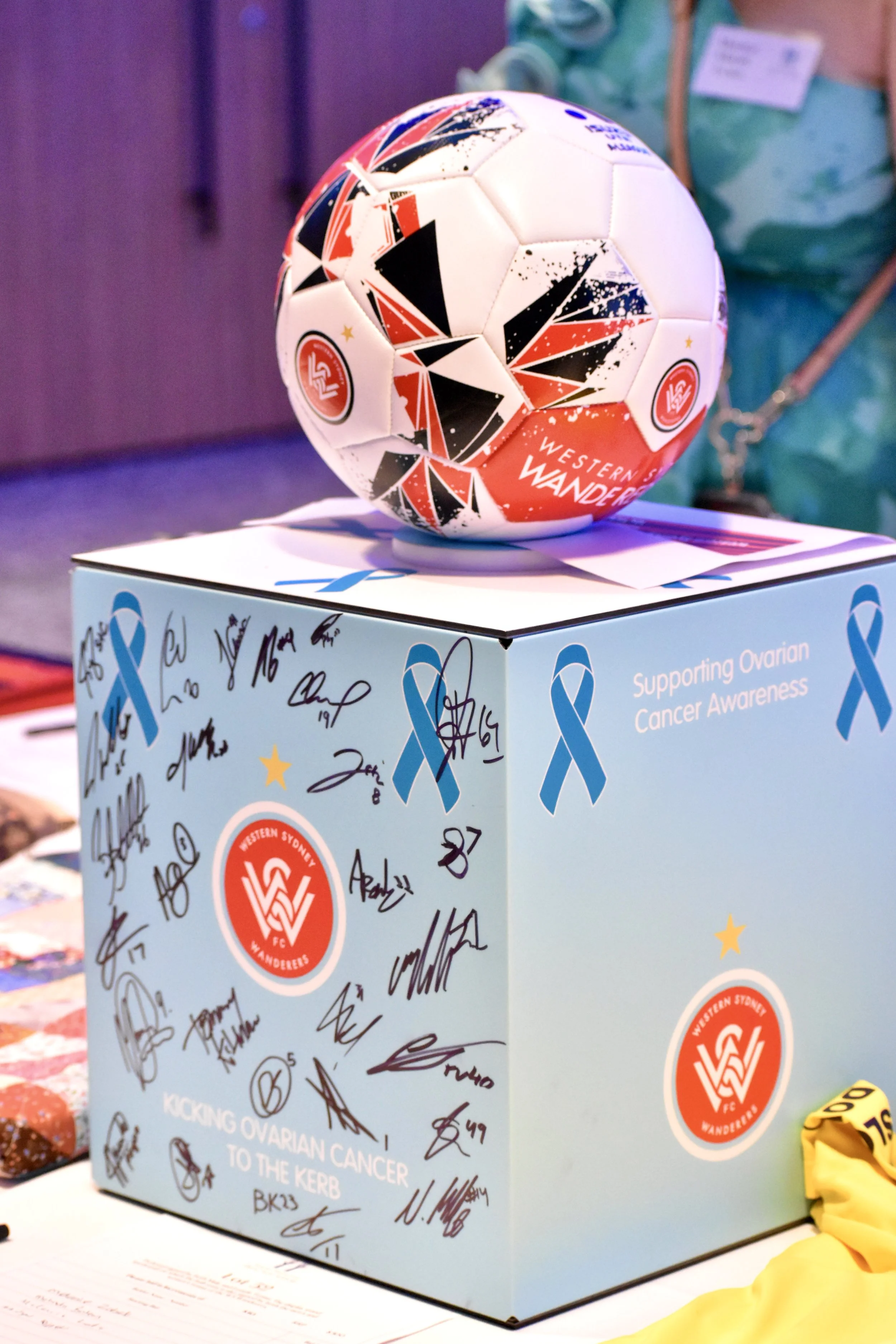 A soccer ball with colorful geometric designs resting on top of a blue donation box with signatures and symbols supporting ovarian cancer awareness for Western Sydney Wanderers FC.