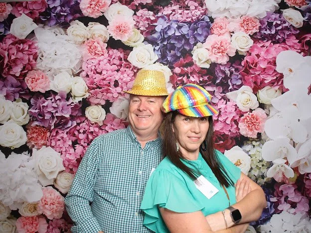 A man and woman standing in front of a floral backdrop, wearing colorful hats. The man wears a gold, glittery hat, and the woman wears a rainbow-striped hat, both smiling and posing.