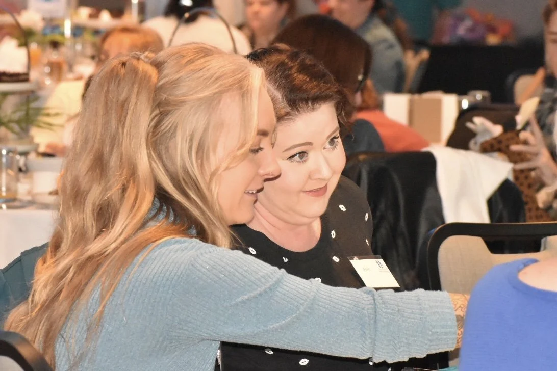 Two women sitting close together, one with blonde hair and the other with dark hair, engaged in a conversation at a conference or event, with other attendees and tables in the background.