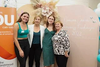 Four women standing together in front of a floral display and a pink backdrop with writing, at an event or celebration.