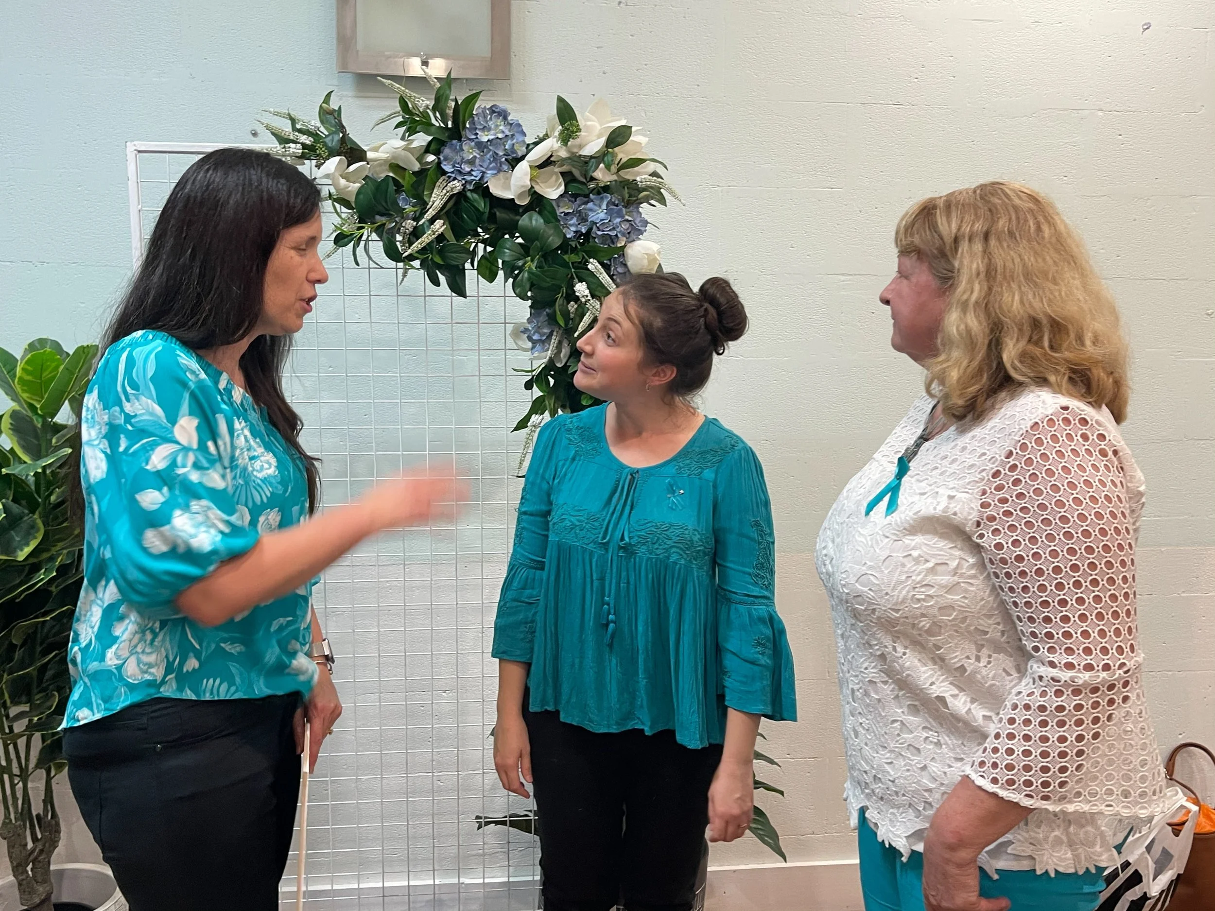Three women standing and talking in front of a floral display with white and blue flowers, on a white wall background.