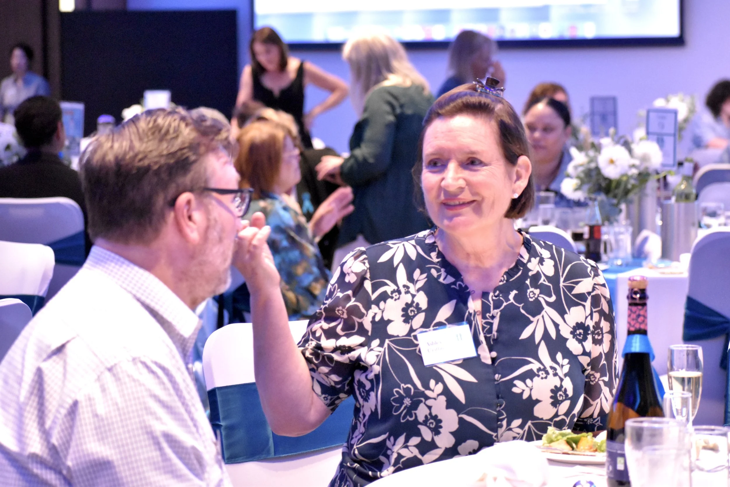 A woman and a man engaged in conversation at a formal event or banquet, with more people seated at tables in the background.