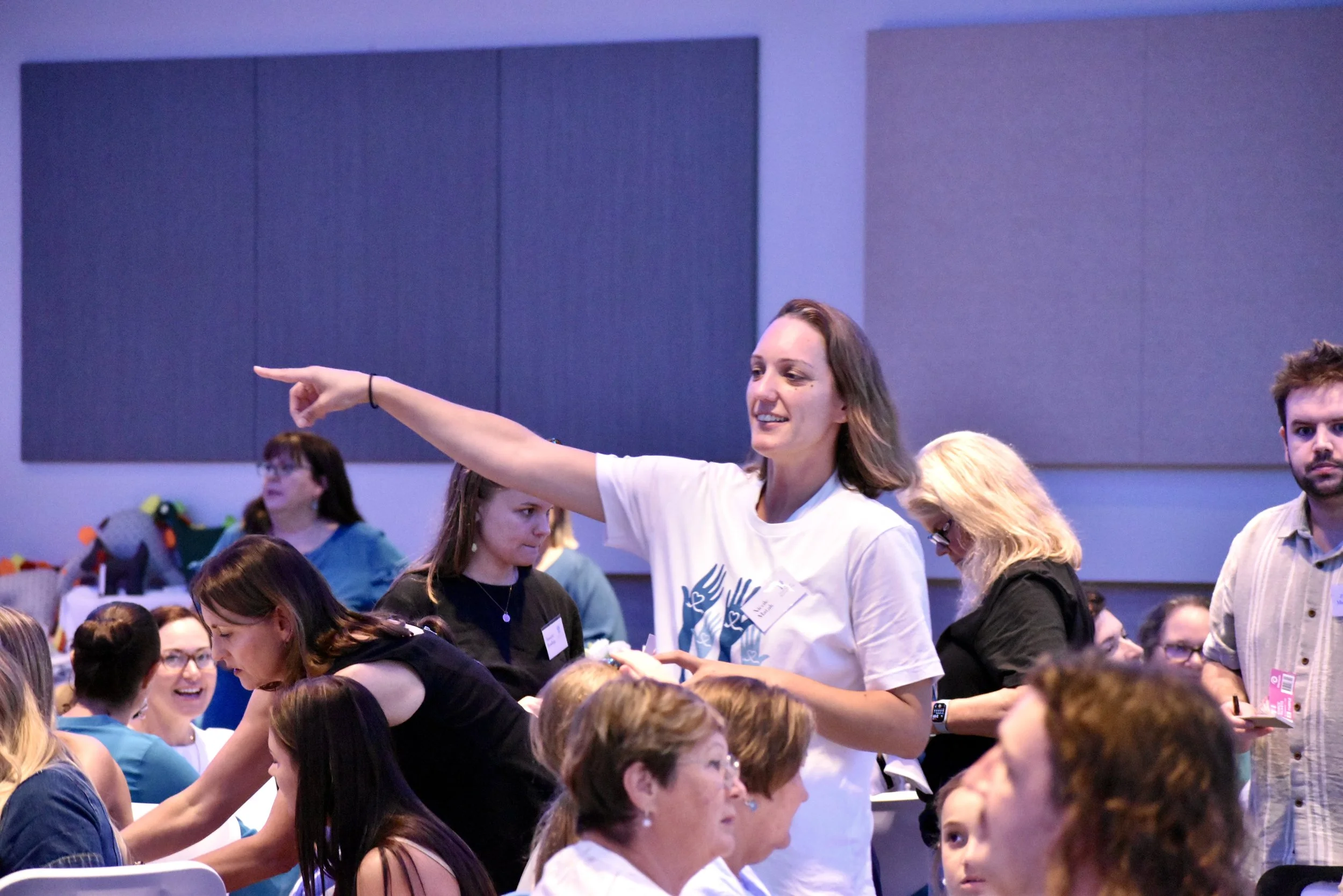 A woman in a white T-shirt is talking to a group of people at a conference or event, gesturing with her arm extended and finger pointing, while others sit and stand around her.