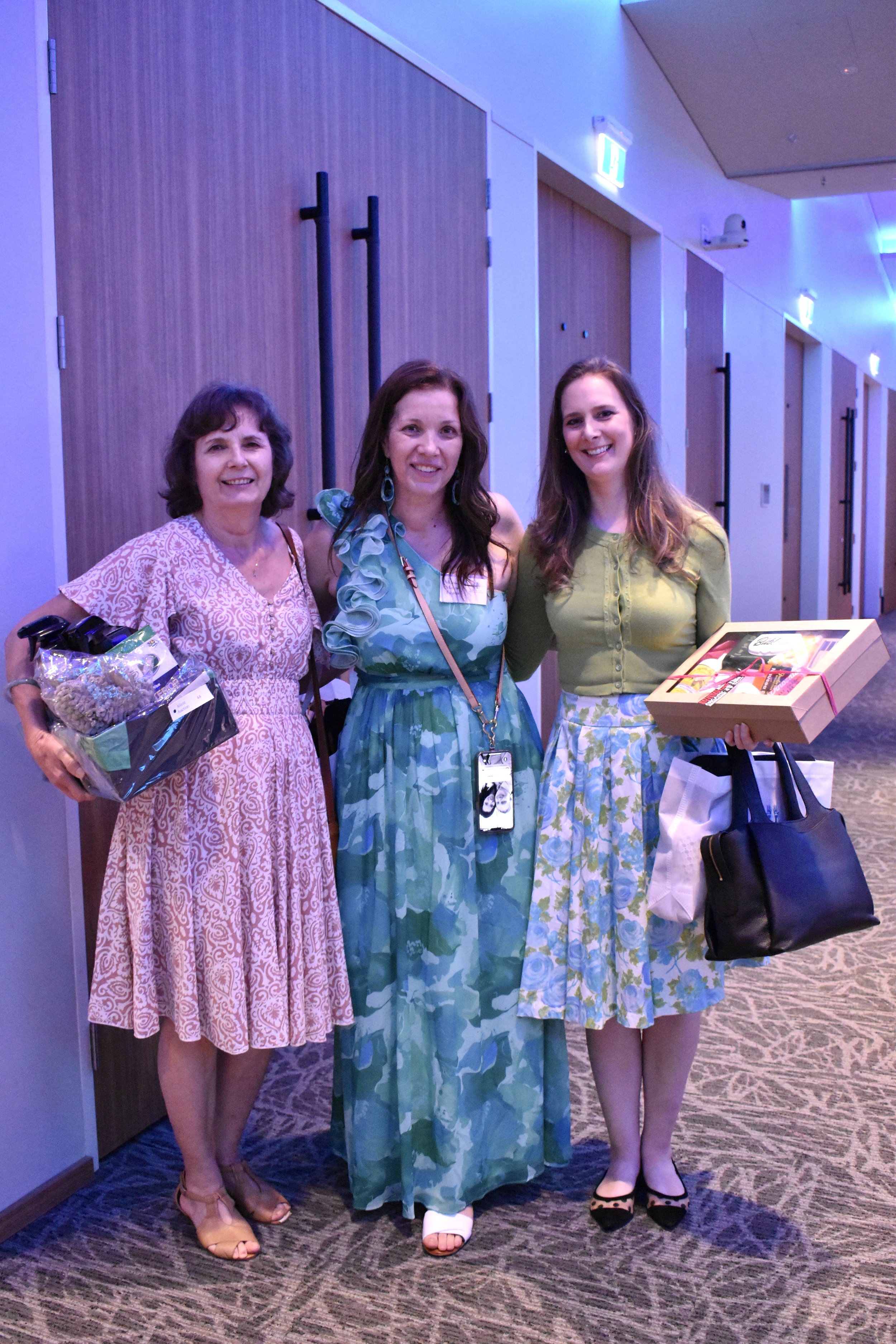 Three women standing in a hallway at an event, smiling, holding gift bags and boxes.