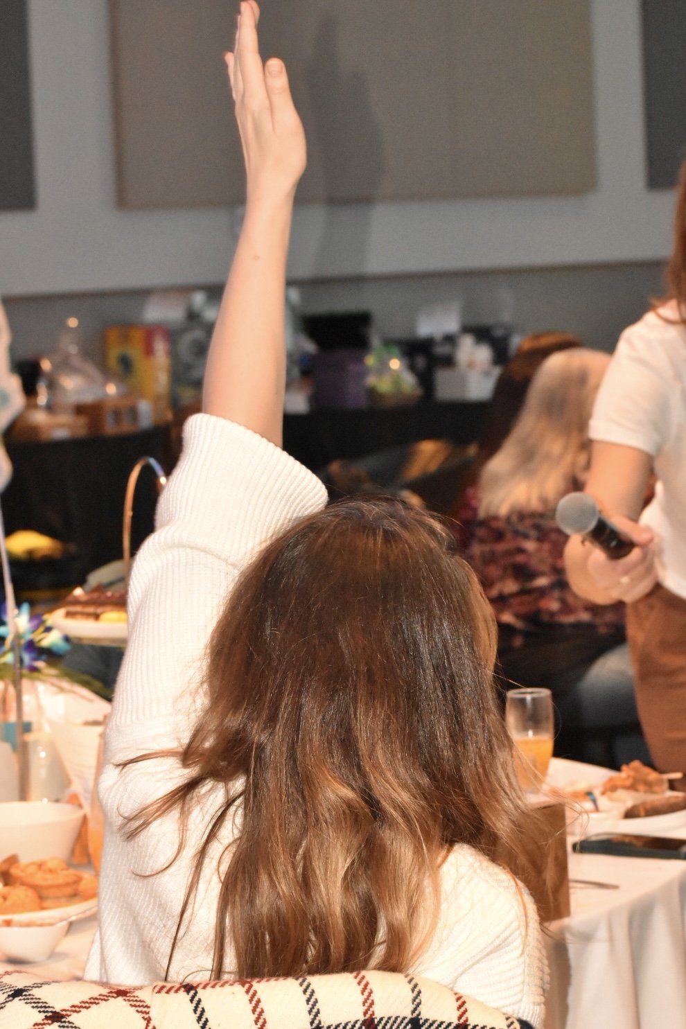 Person with brown hair raising their hand at a gathering or meeting, with food and drinks on the table, and a woman holding a microphone on the right side.
