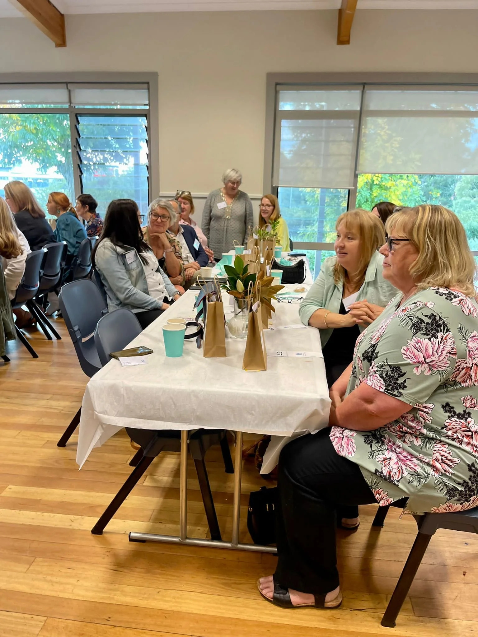 A group of women gathered around a table with brown gift bags, plants, and cups, in a bright room with large windows and wooden floors.