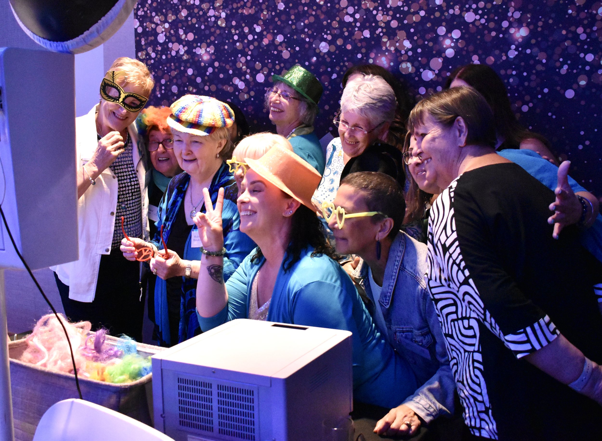 A group of women, some wearing colorful and fun accessories like hats and glasses, gather around a photo booth, smiling and posing for a picture with a purple and pink bokeh background.