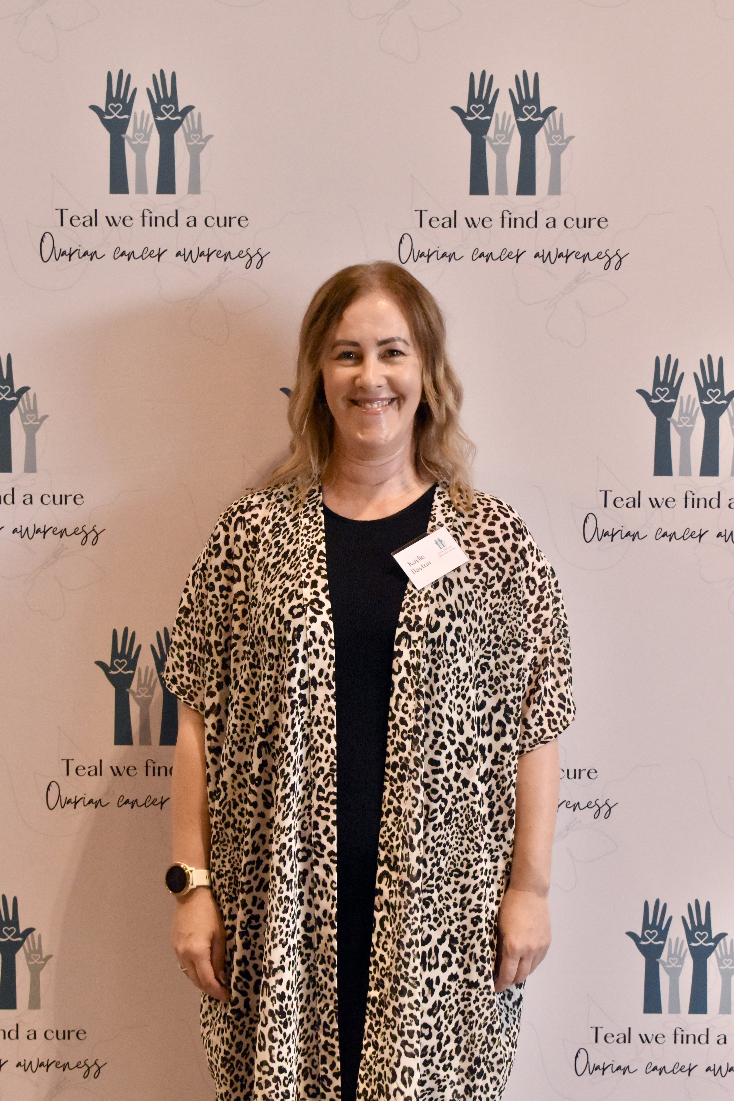 Woman in front of ovarian cancer awareness backdrop, wearing a leopard print kimono style jacket and black top, with a name tag that reads 'Katie Benson'.