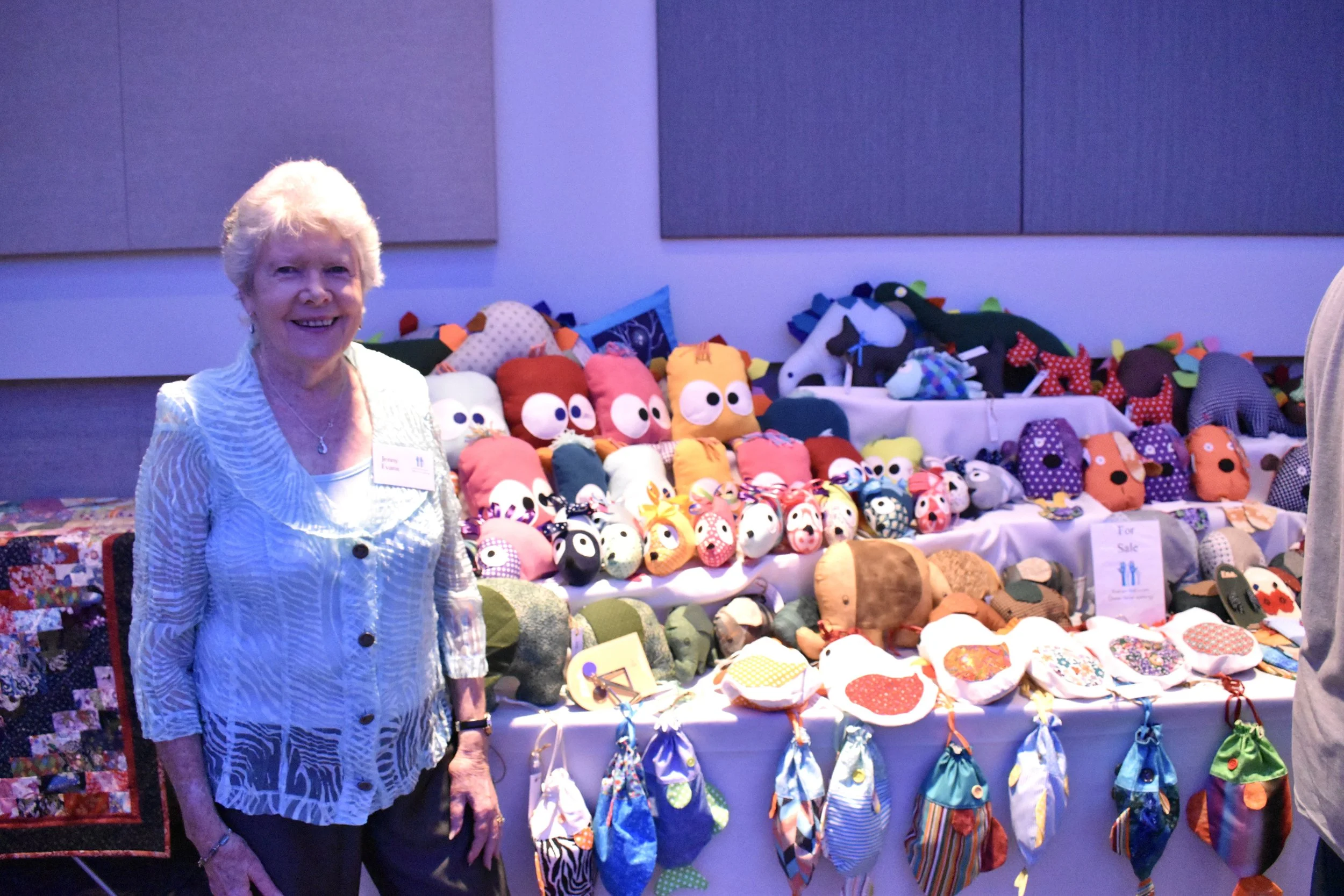 A woman standing in front of a table filled with handmade fabric stuffed animals and toys at an indoor craft fair.