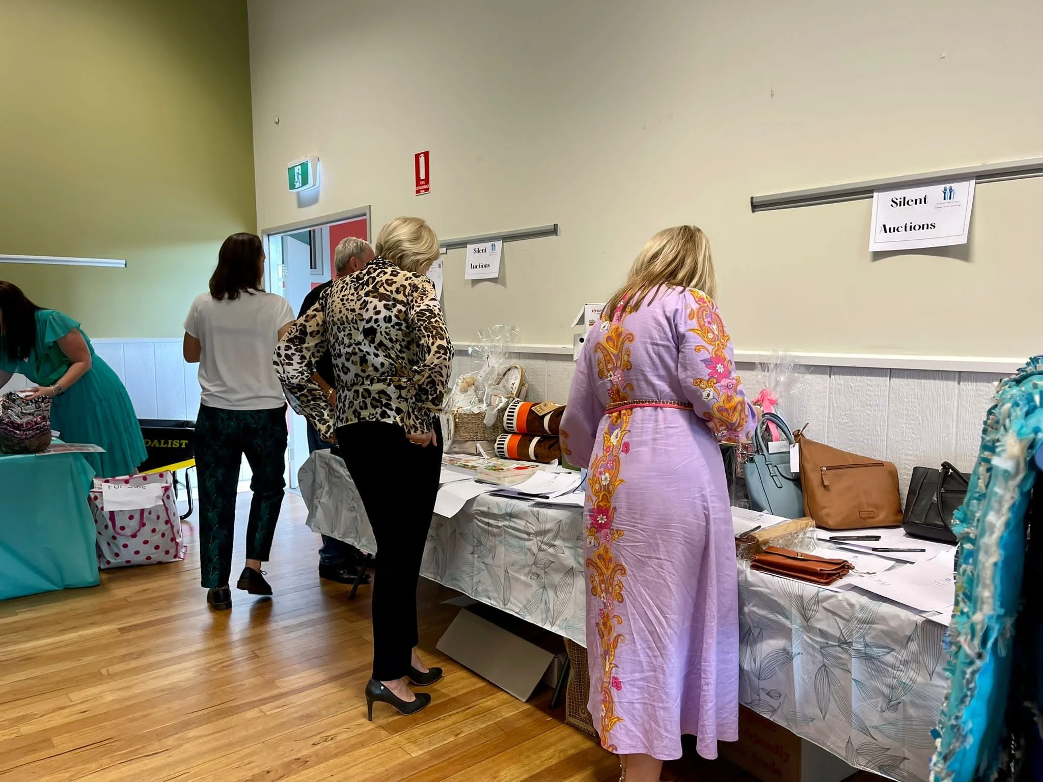 People shopping at a silent auction in a room with tables displaying items and bags.