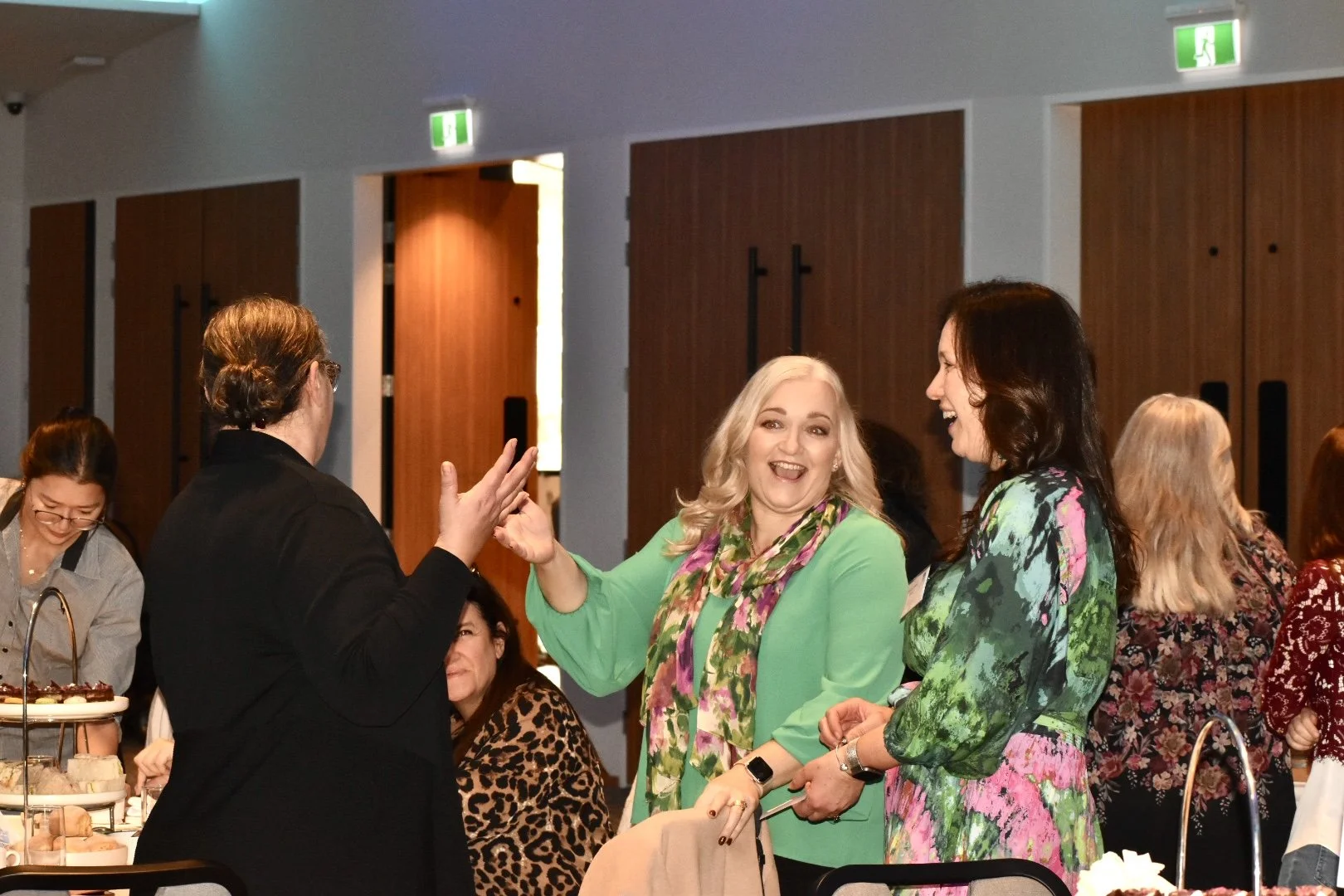 Two women smiling and high-fiving each other at a social gathering, with other women in the background. One woman has blonde hair and is wearing a green dress with a floral scarf; the other has dark hair and is wearing a colorful patterned dress.