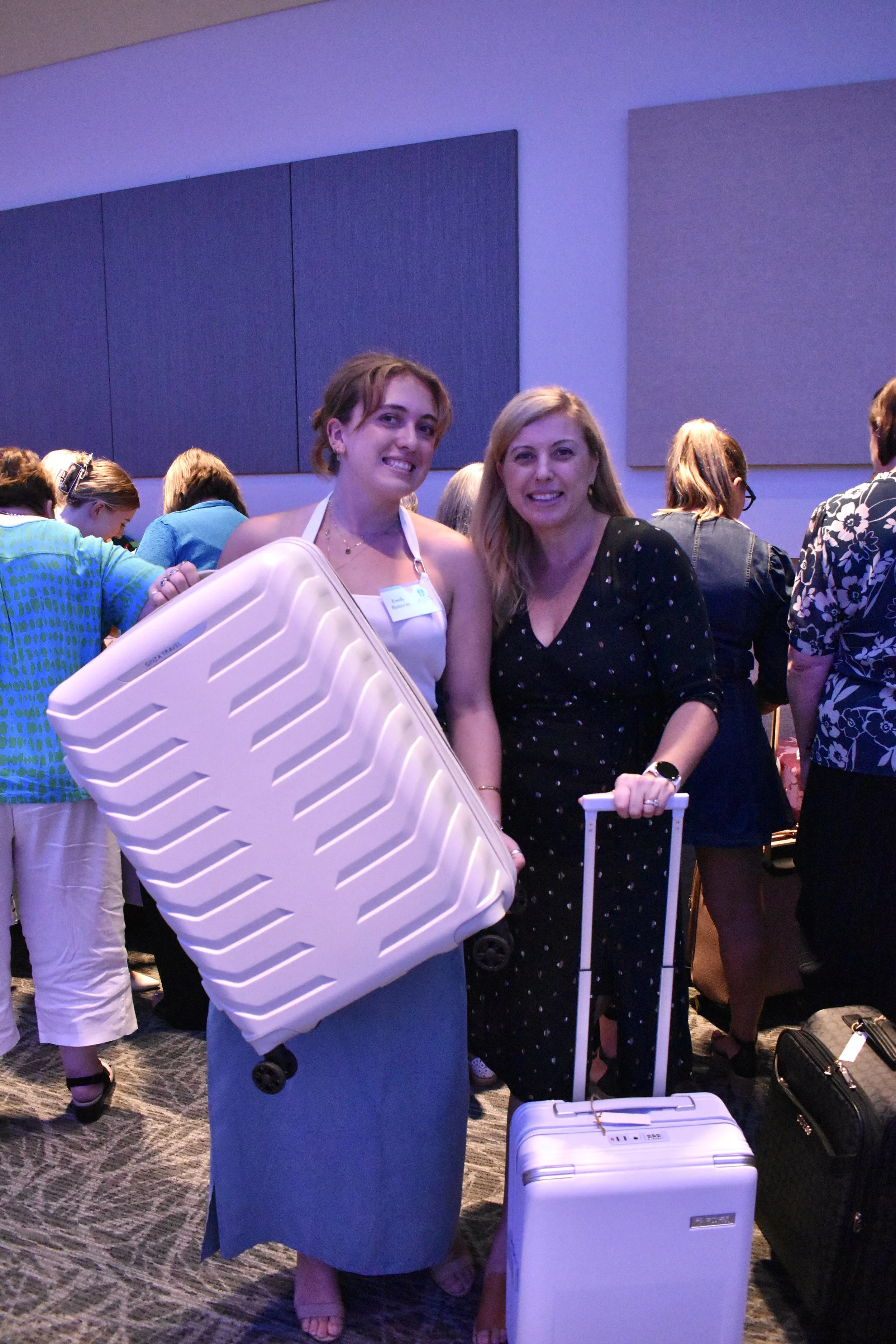 Two women standing together at an event, one holding a large white suitcase and the other holding a smaller white carry-on suitcase, with many people in the background.