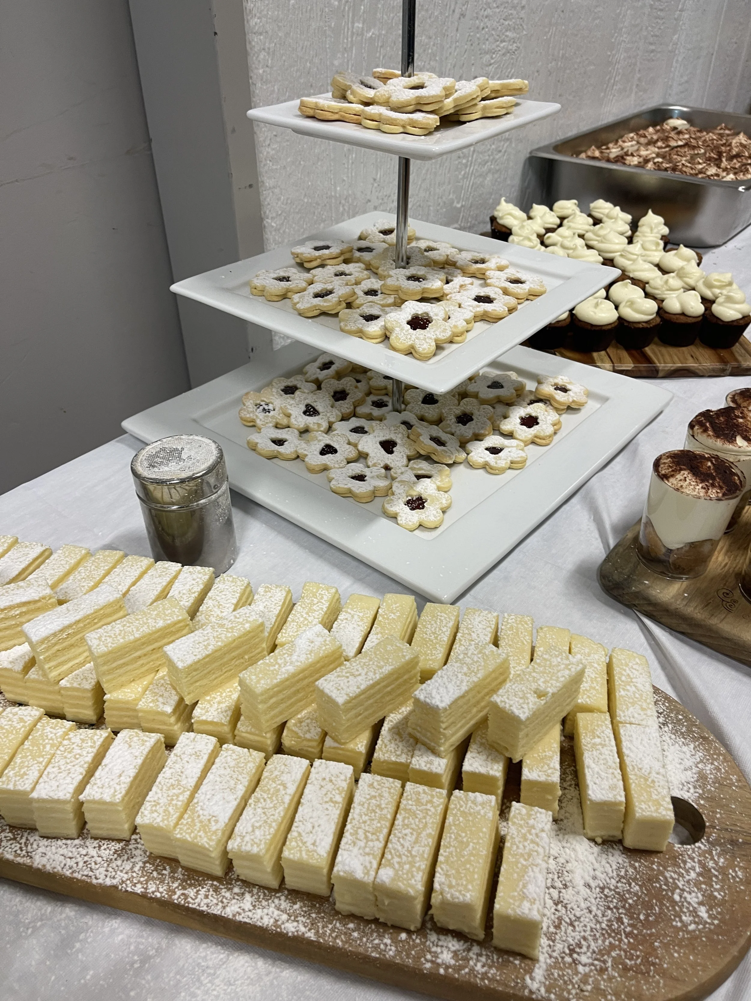 Assorted desserts including layered cake slices, flower-shaped cookies with powdered sugar and jam centers, cupcakes with white frosting, and other sweets on a table.