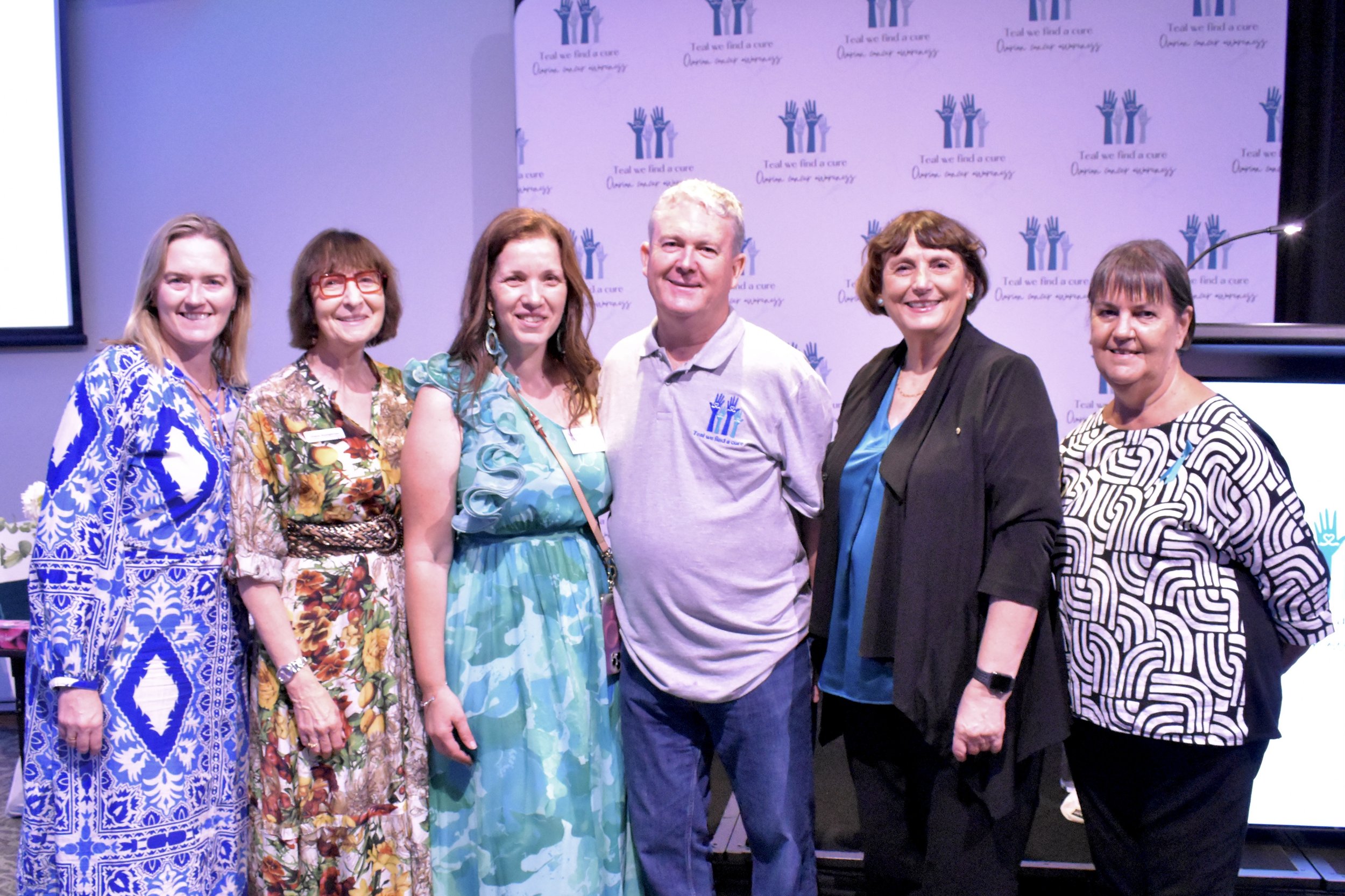 Group of seven people posing together at an event, standing in front of a backdrop with a logo and text that reads 'Tear we find a cure, Ovarian cancer awareness'.