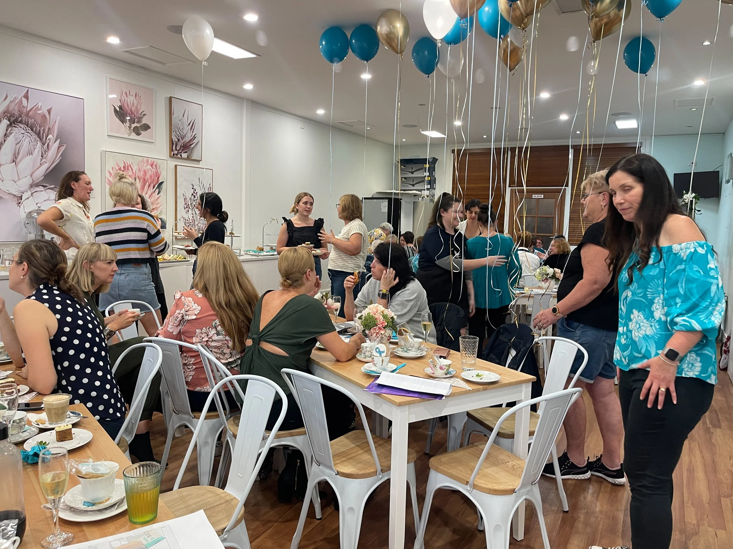 A group of women gathered for a celebration in a decorated indoor space with balloons, flowers, and a long table with food and drinks.