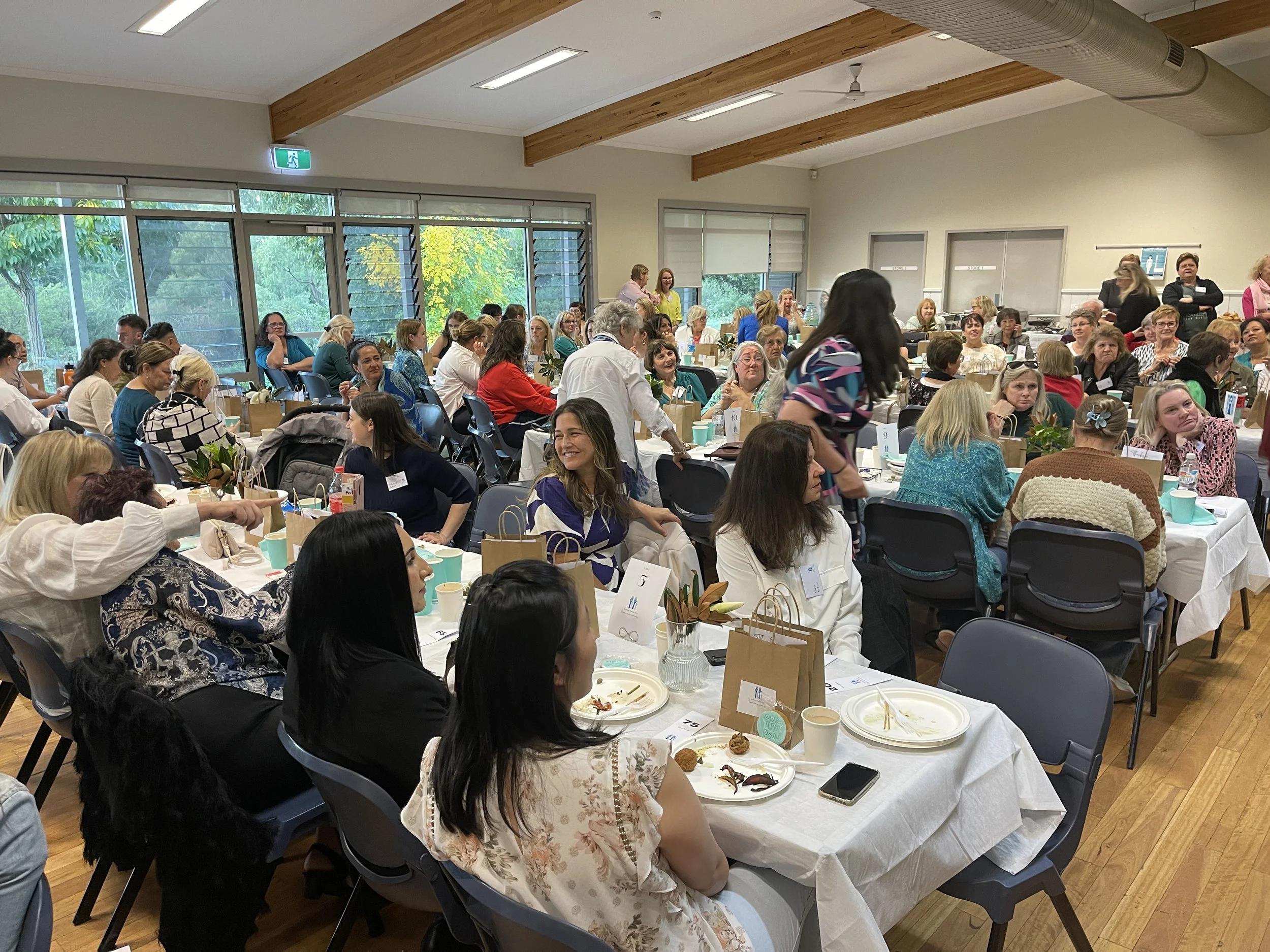 A large gathering of women in a well-lit event space, seated at decorated tables with food and drinks, engaging in conversations and activities during a social or professional event.