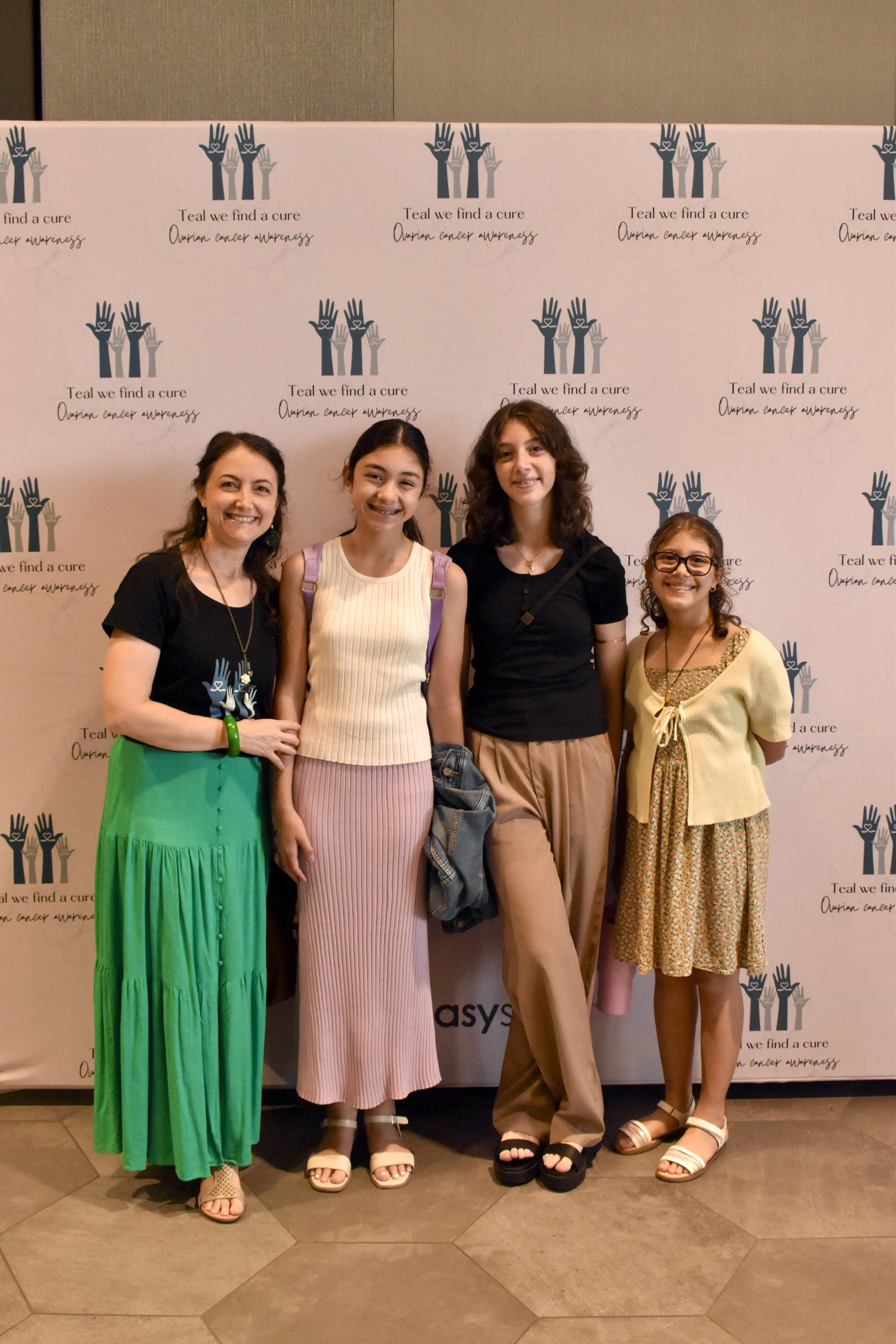 Four women standing together at an awareness event, with a pink backdrop featuring the logo and slogan for ovarian cancer awareness.