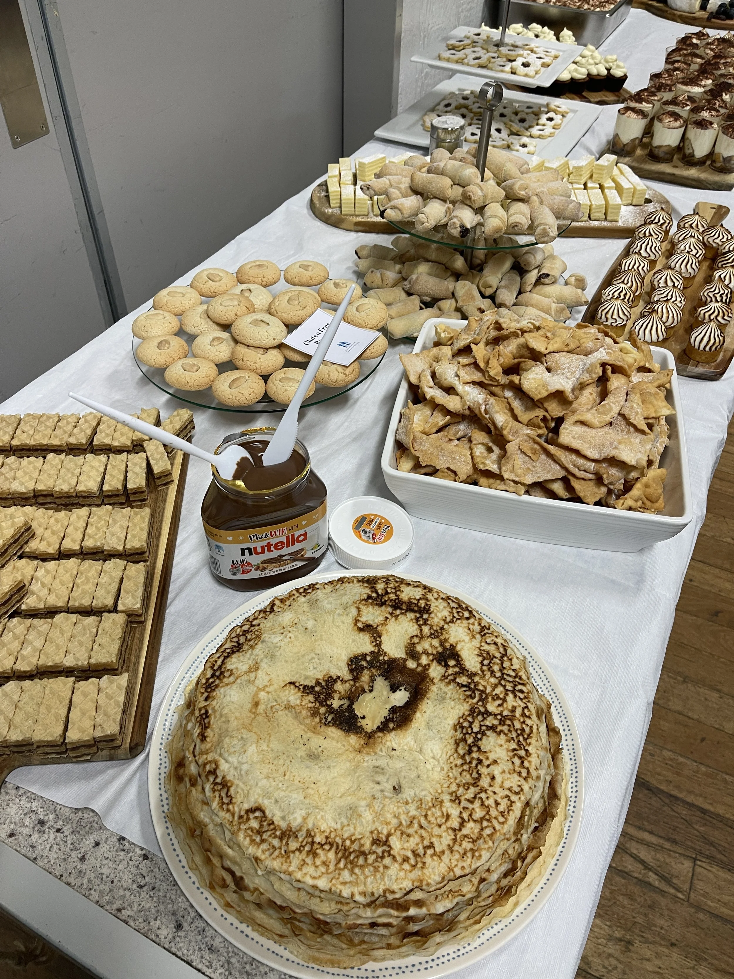 A dessert table with a large crepe cake, cookies, cannoli, cookies and cream cupcakes, piped cookies, and various other sweet treats, along with a jar of Nutella.