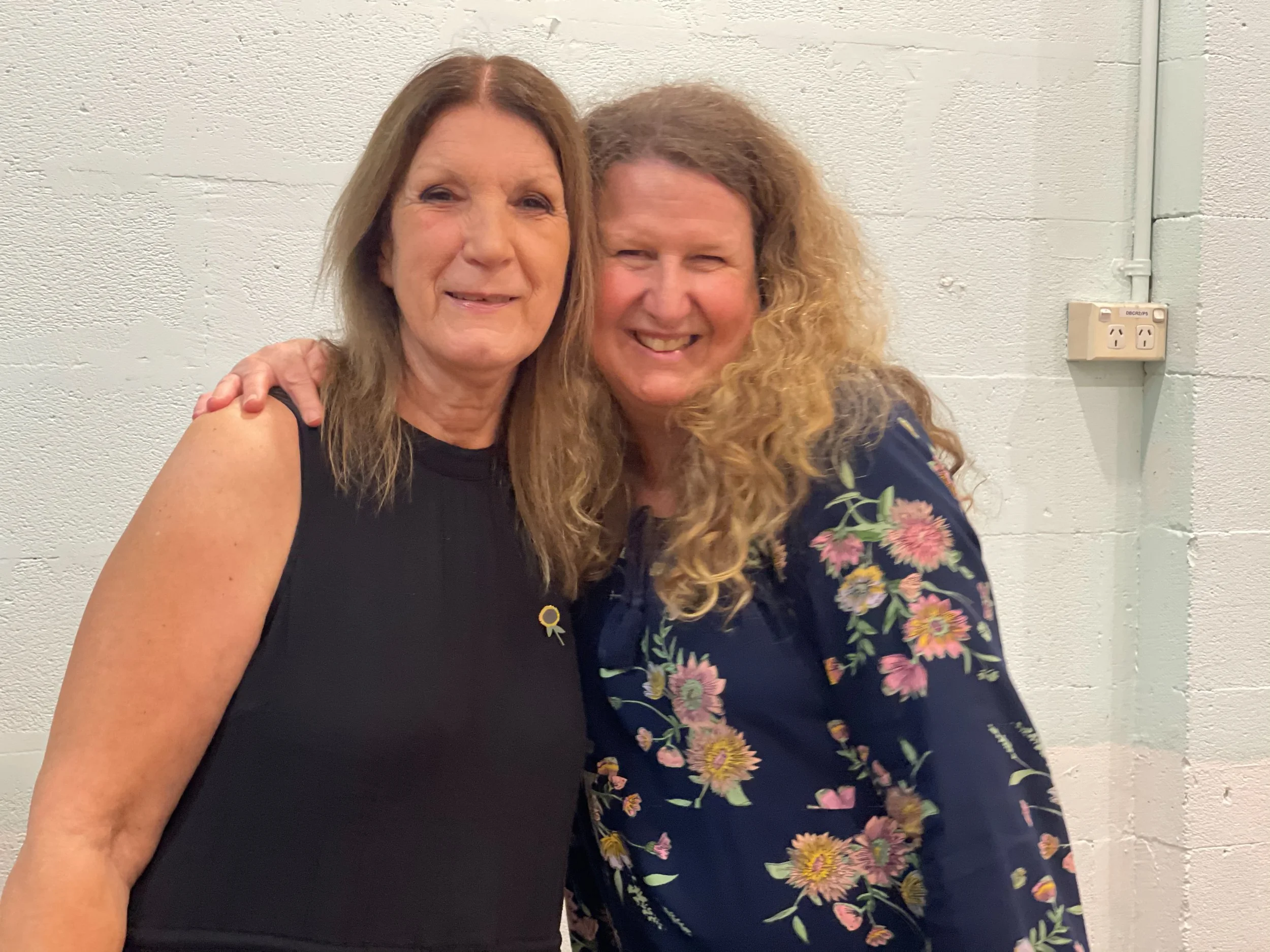 Two women standing close together, smiling, with one placing her arm on the other's shoulder, against a plain white wall background.