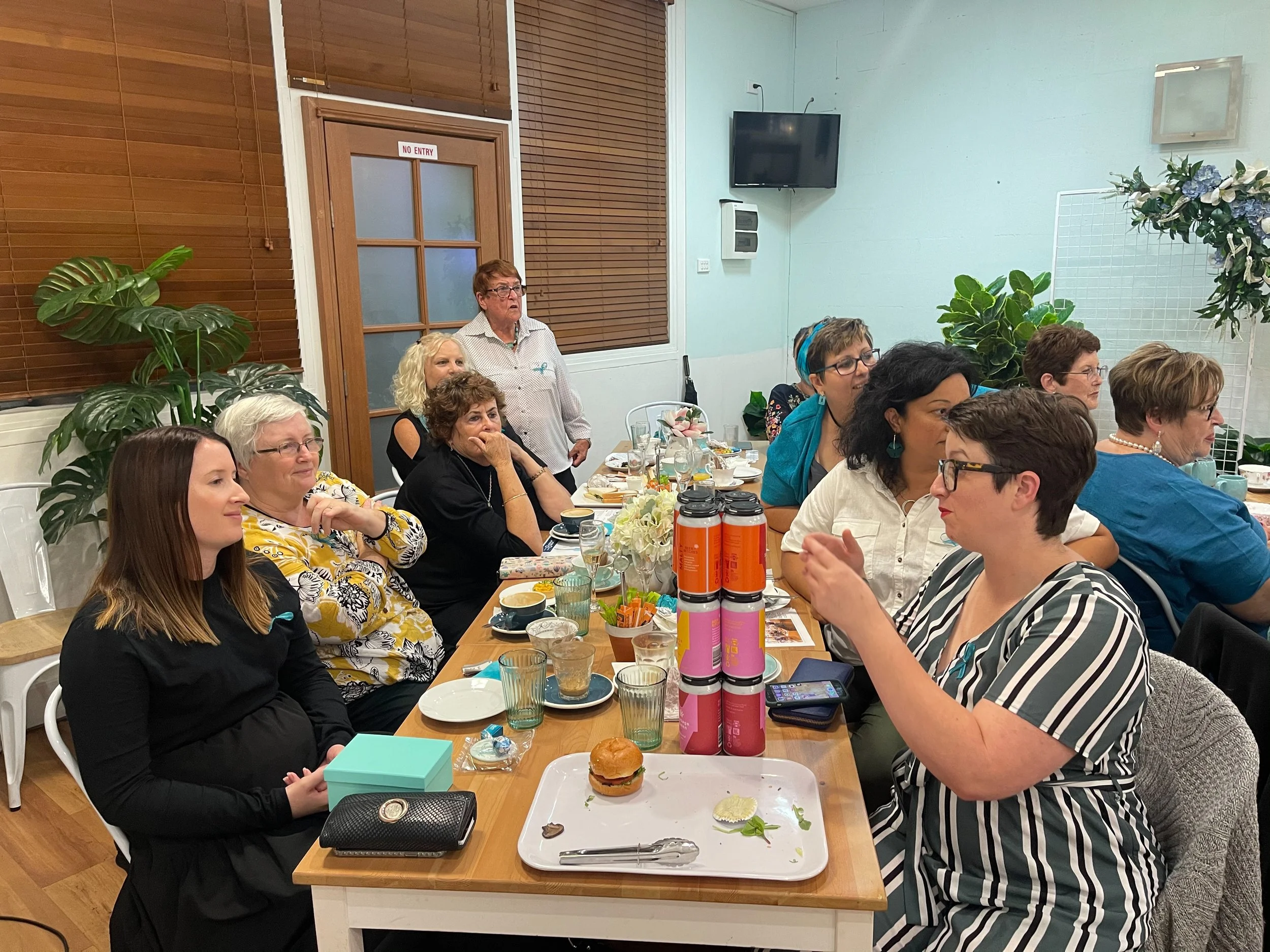 Group of women seated at a dinner table in a room decorated with plants and flowers, engaged in conversation, with some people listening attentively.