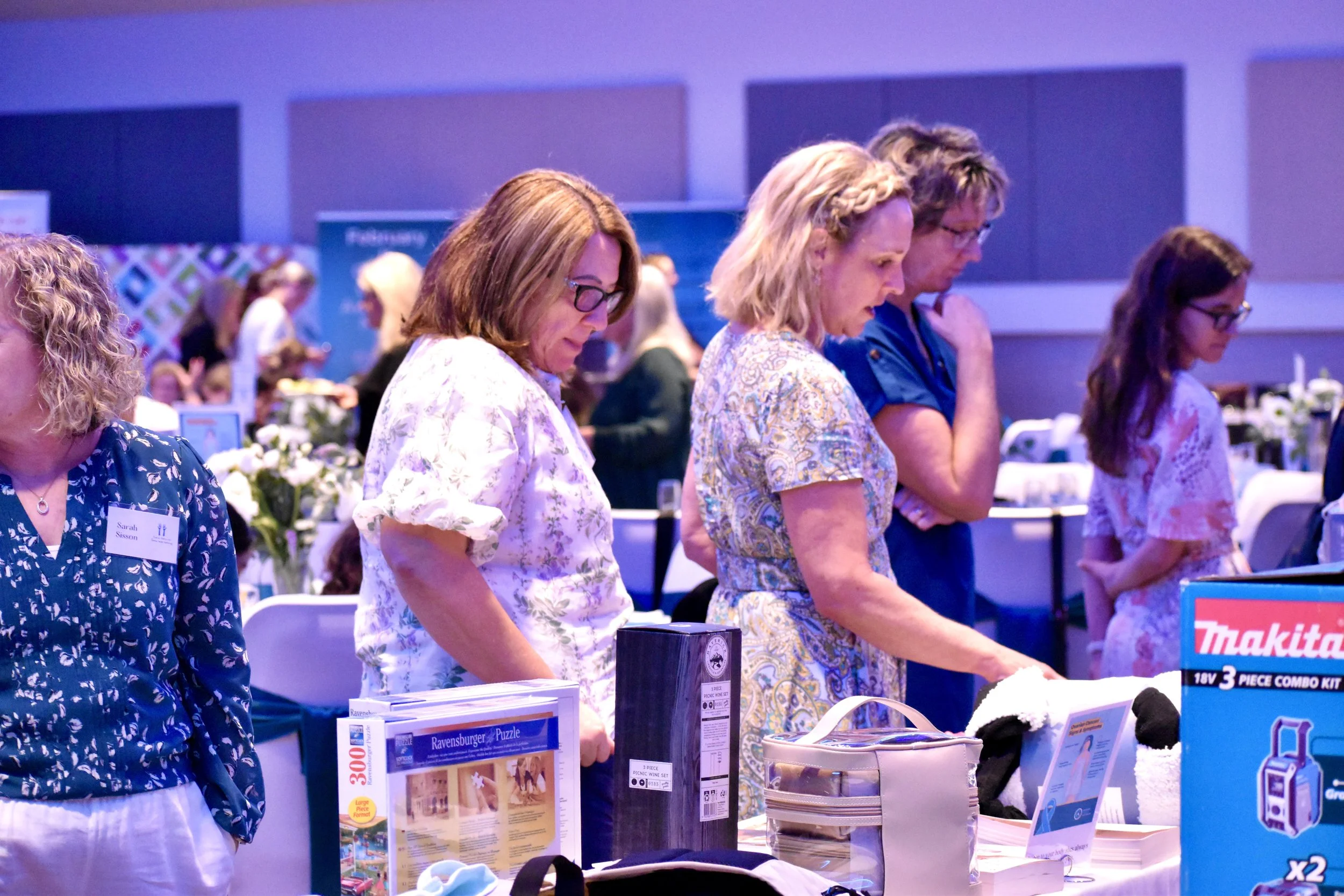 Four women standing at a table looking down at items, with one woman wearing glasses and a floral shirt, and others in patterned dresses, in an indoor event space with tables and people in the background.
