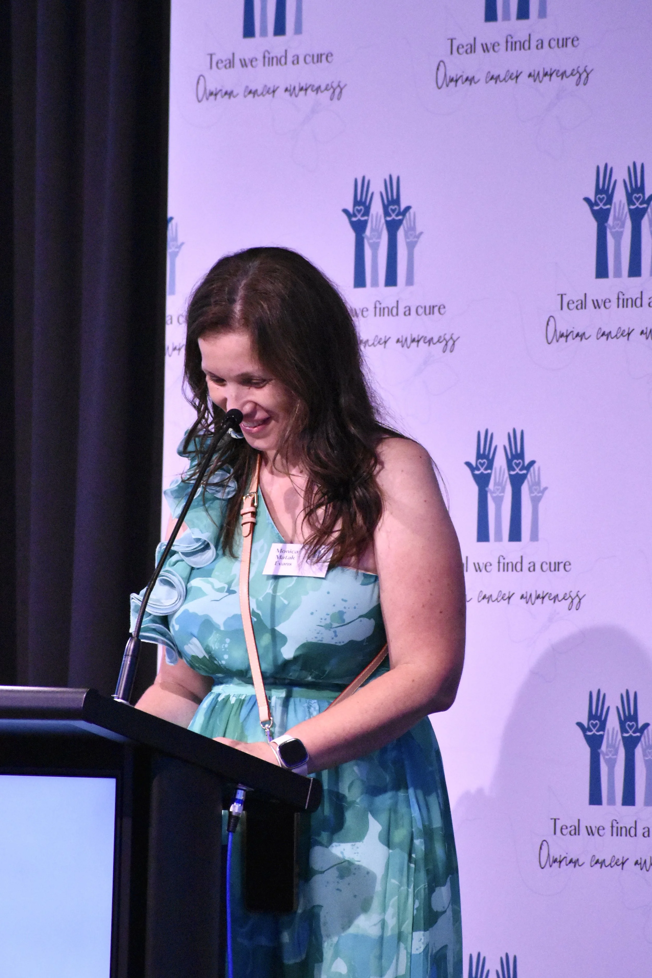 A woman with long brown hair standing at a podium, smiling while speaking into a microphone, wearing a green and blue patterned dress with a flower detail on her shoulder, and a smartwatch on her left wrist, at an event with a backdrop that has blue 