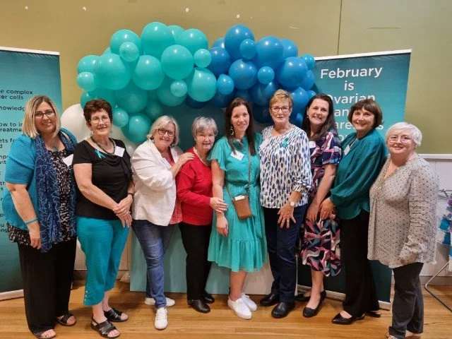 Group of nine women standing together indoors in front of teal and blue balloons and informational banners.